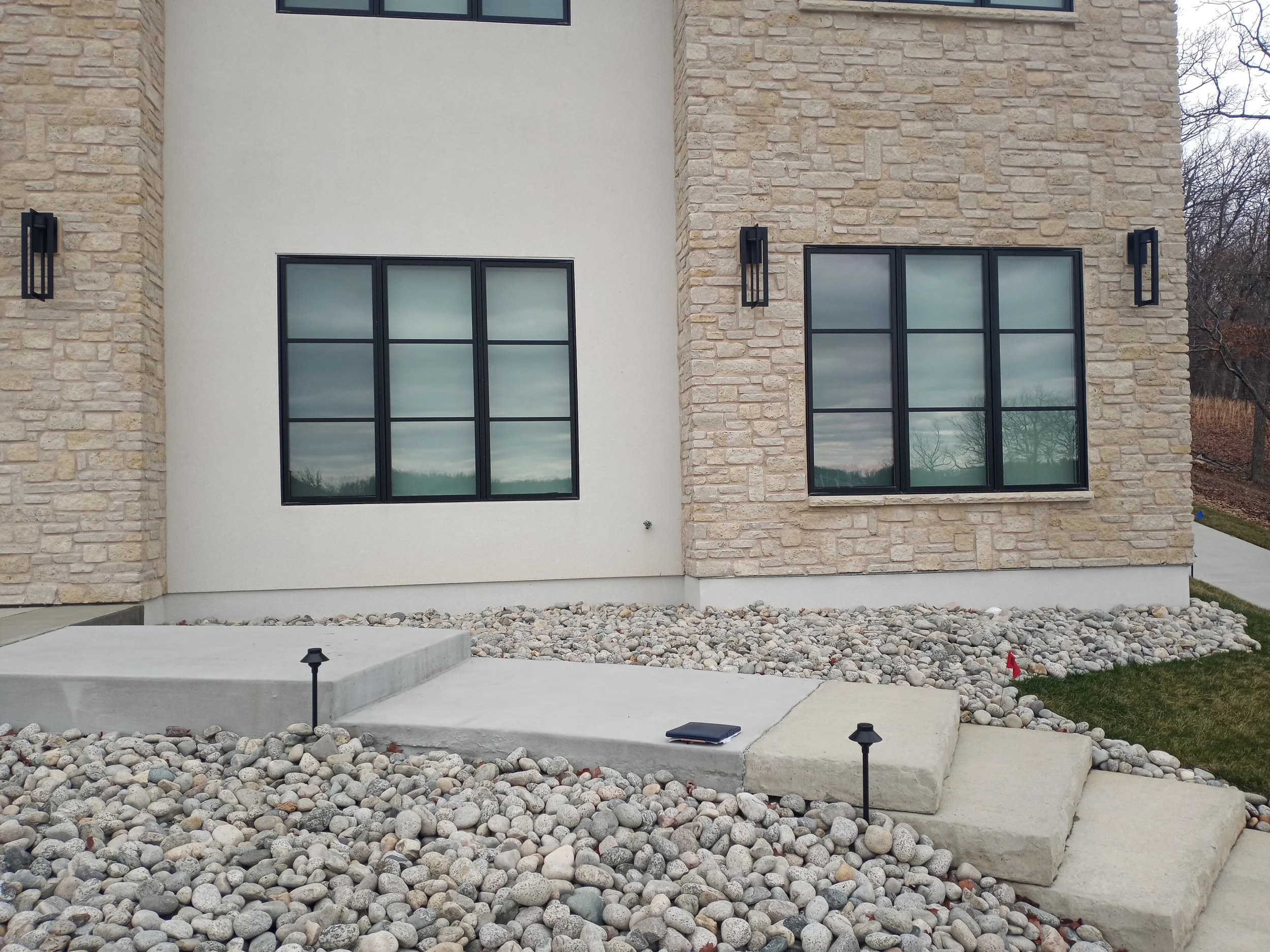 Photo of the front exterior of a modern house with beige stone and white stucco walls, featuring large black-framed windows and outdoor lighting fixtures, with a pathway and rock landscape.