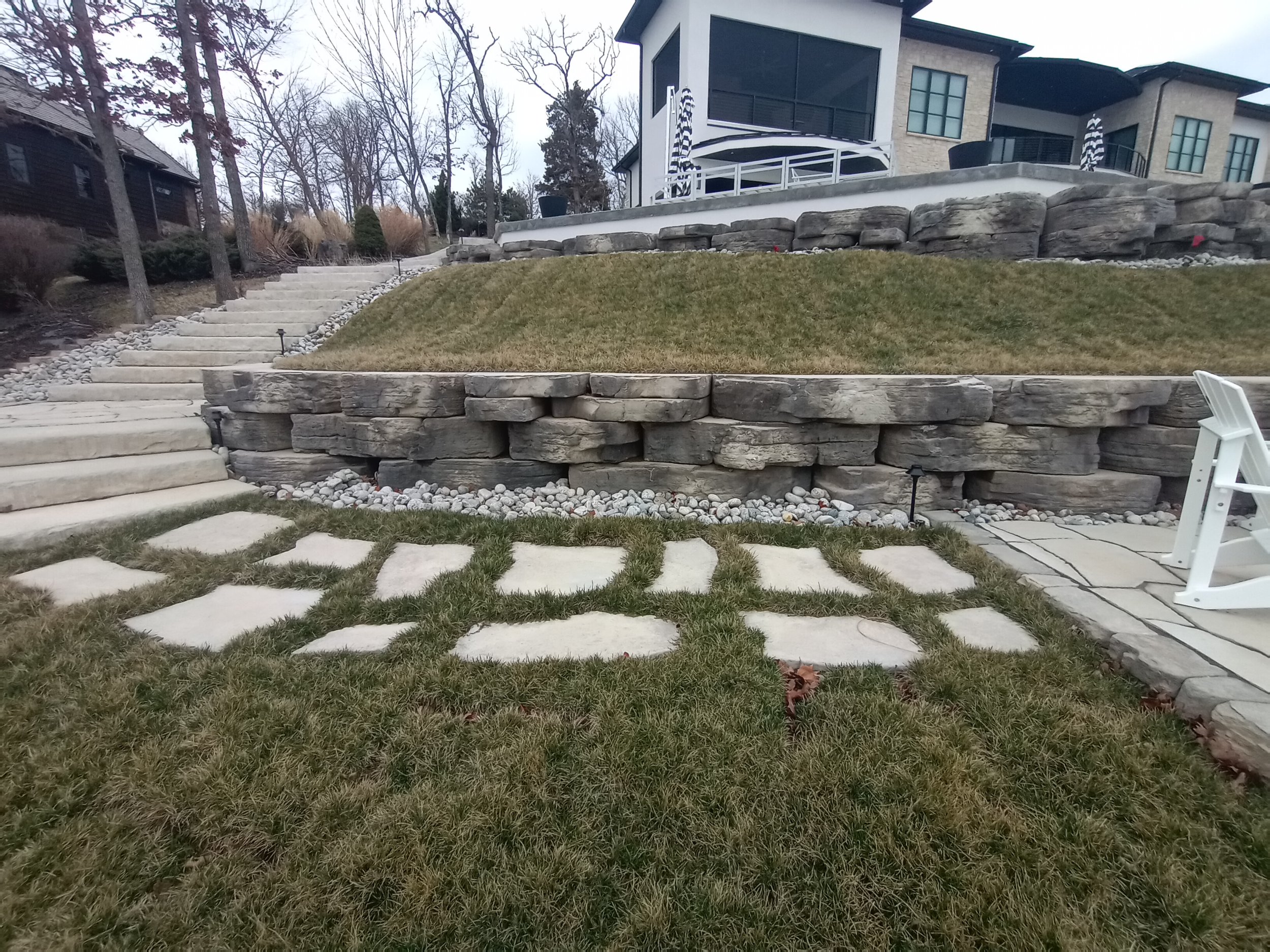 A landscaped backyard with stone steps, a Grass lawn, a tiered stone retaining wall, decorative rocks, and outdoor furniture.