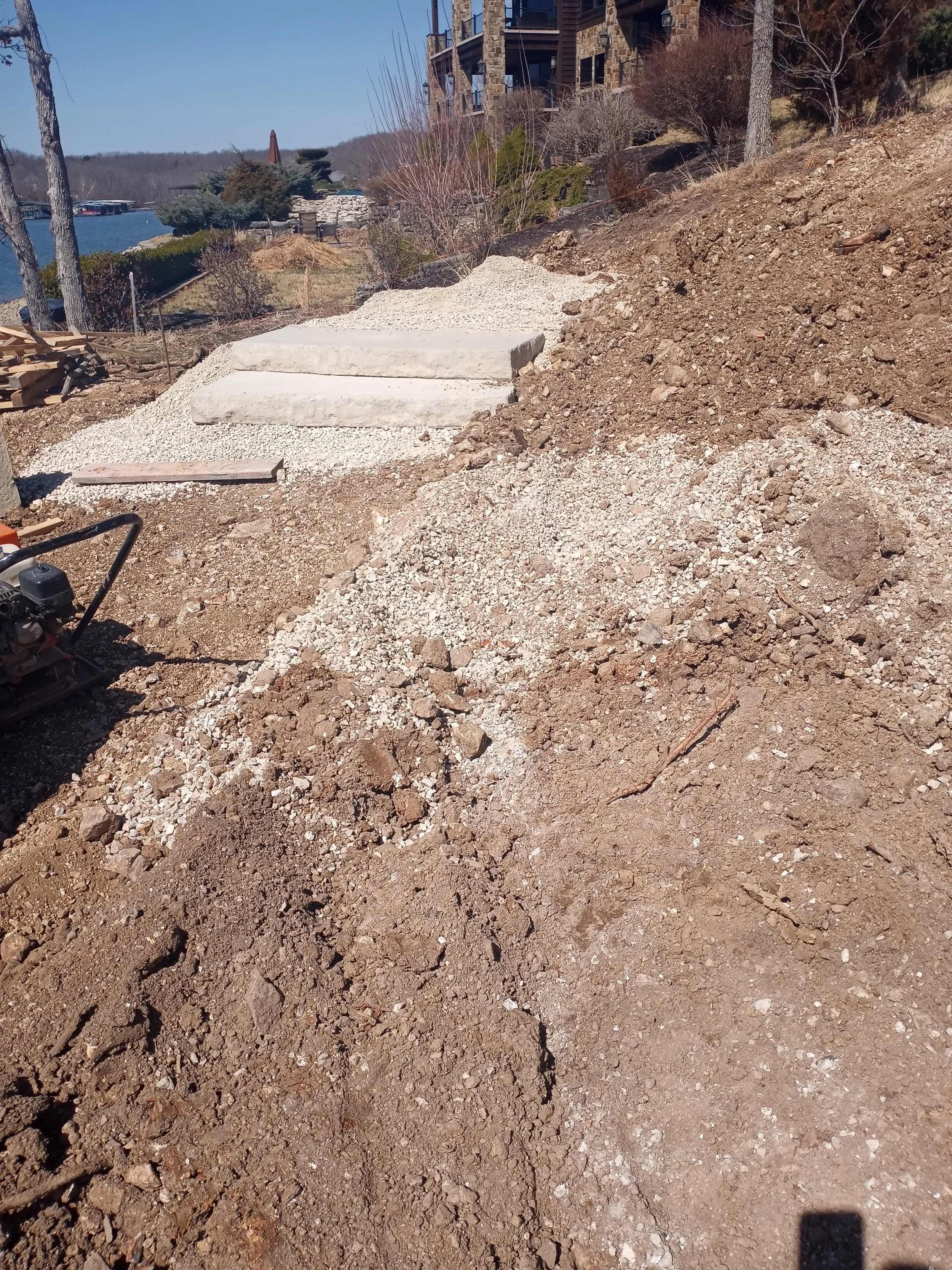Construction site with soil and gravel, concrete steps, and a nearby body of water in the background.