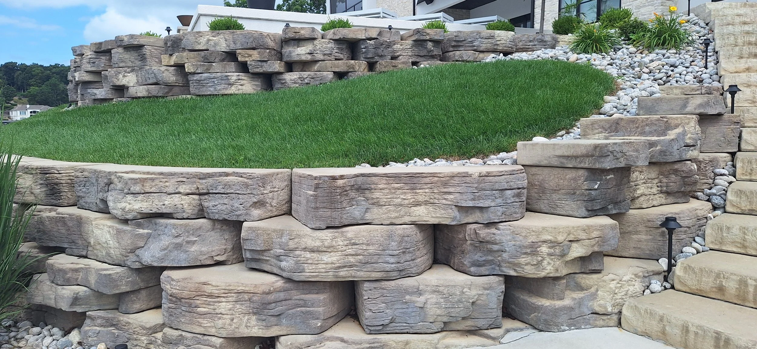 Landscape of a tiered garden with layered large stone walls, green grass, small white decorative rocks, and plants with yellow flowers. Modern house visible in the background.