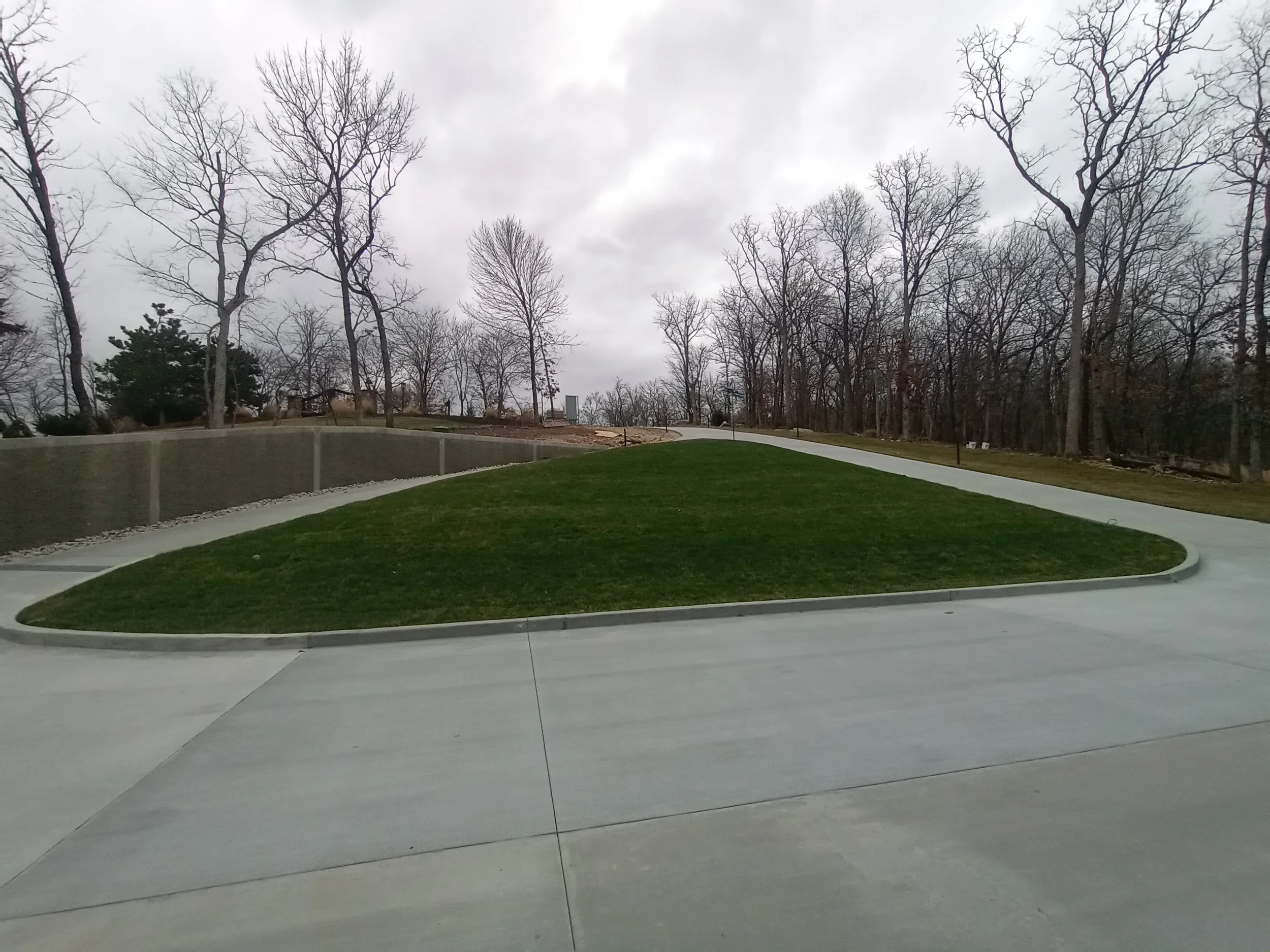 A small grassy hill with a sidewalk around it, with bare trees in the background and a cloudy sky.