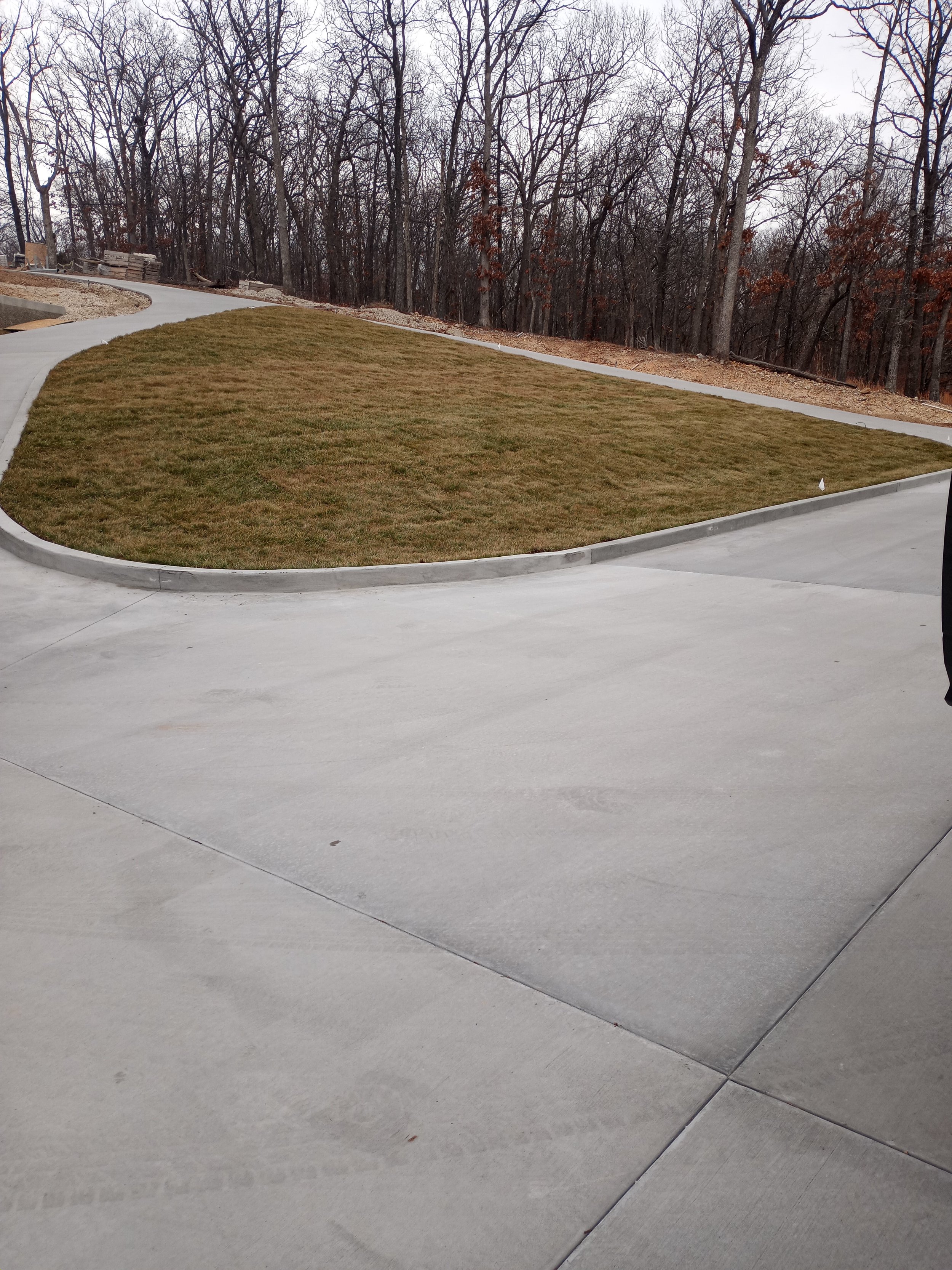 A curved concrete driveway with a grass medallion in the center, surrounded by leafless trees under a cloudy sky.