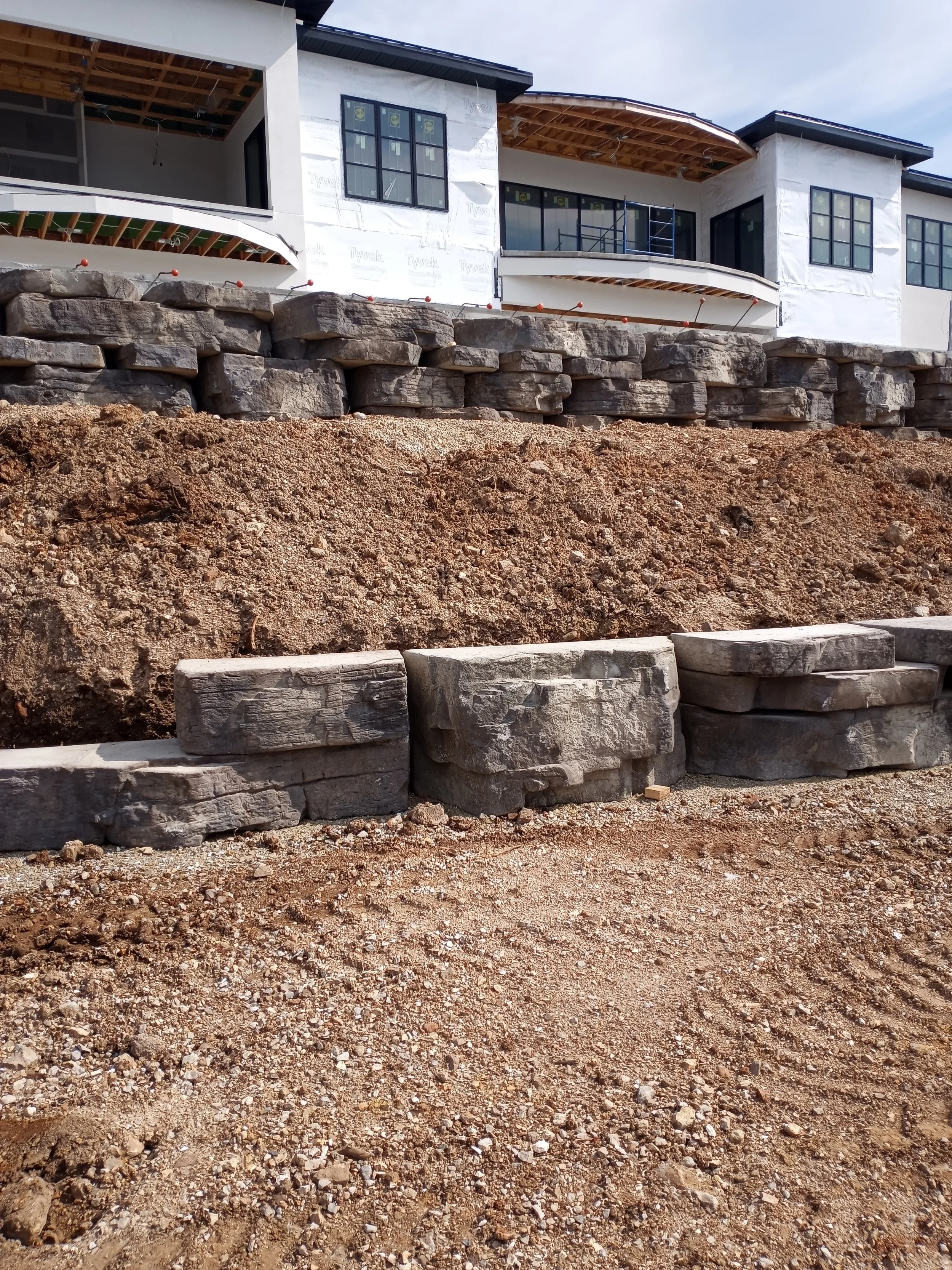 Construction site with retaining wall of large stones and an unfinished white house with multiple windows, some scaffolding, and exposed roof framing.