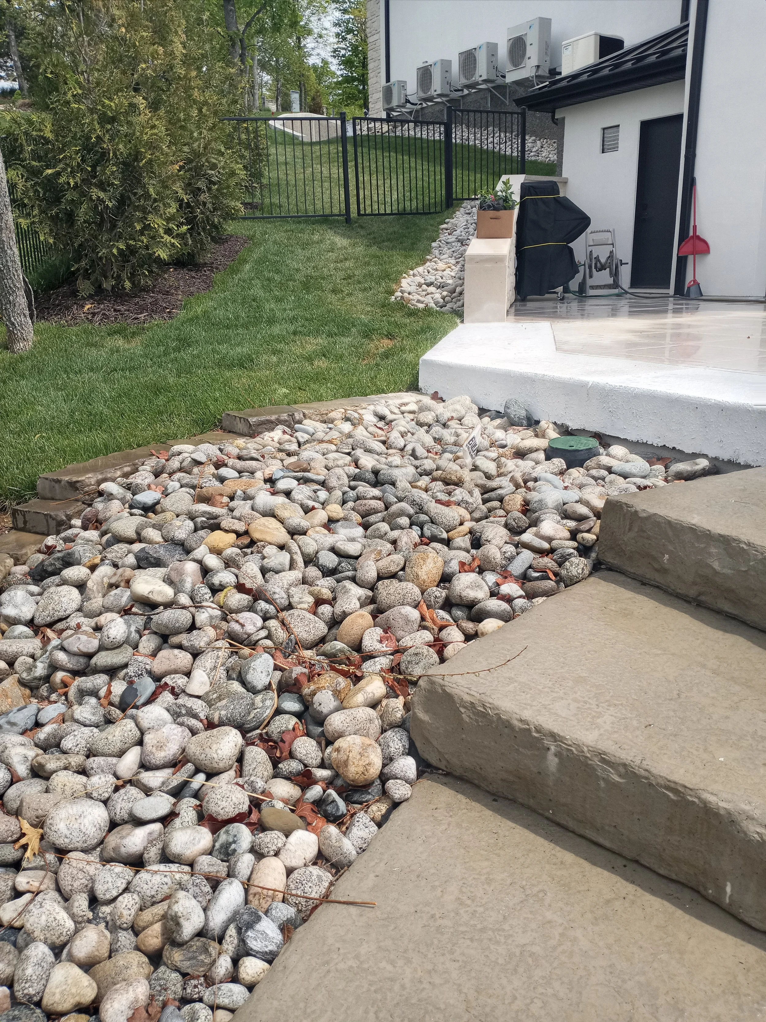A backyard patio area with a white concrete porch, gravel and stone landscaping, a grassy lawn, a black metal fence, and a building with multiple air conditioning units mounted on the exterior wall.