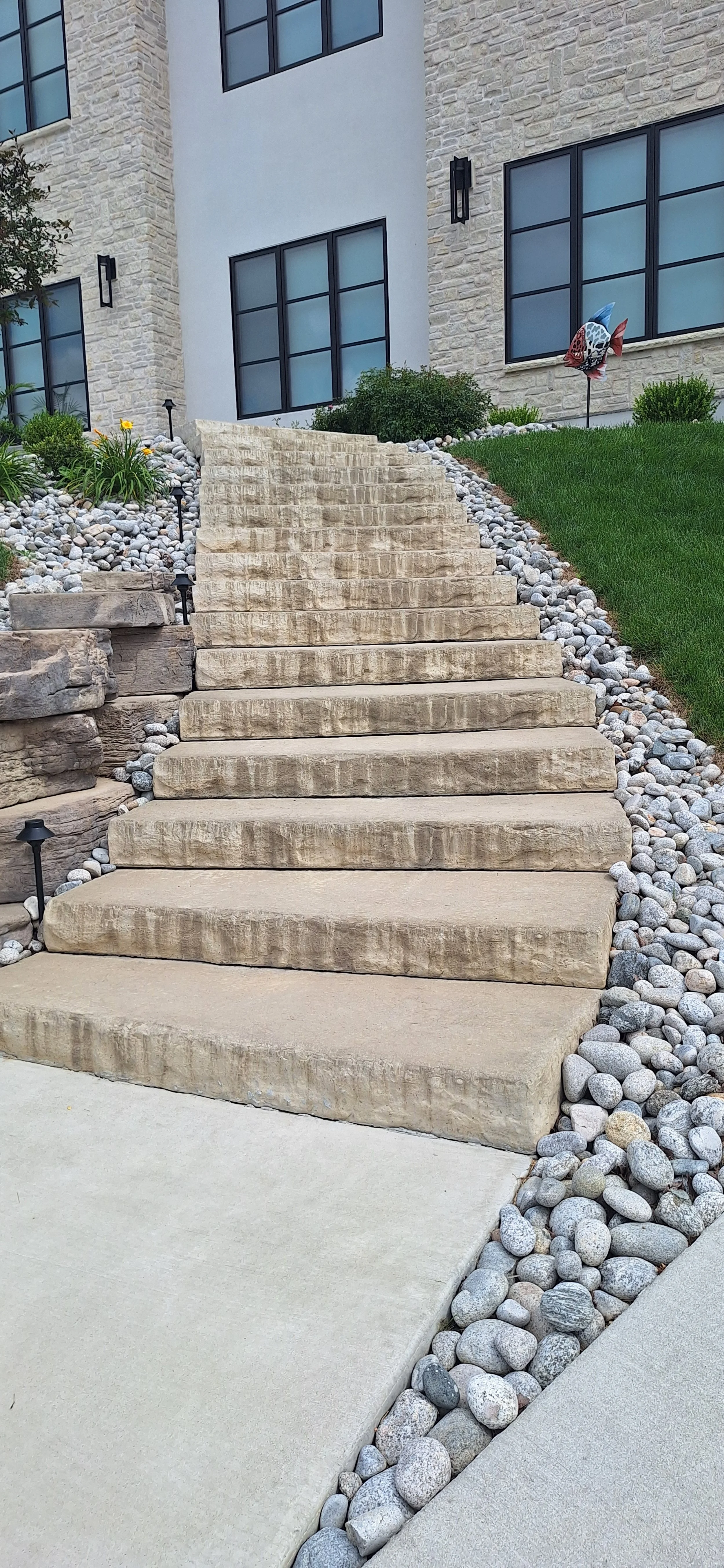 Concrete stairs leading up to a modern residential building with large black framed windows, a stone and siding exterior, surrounded by landscaped greenery, rocks, and a small decorative fish sculpture.