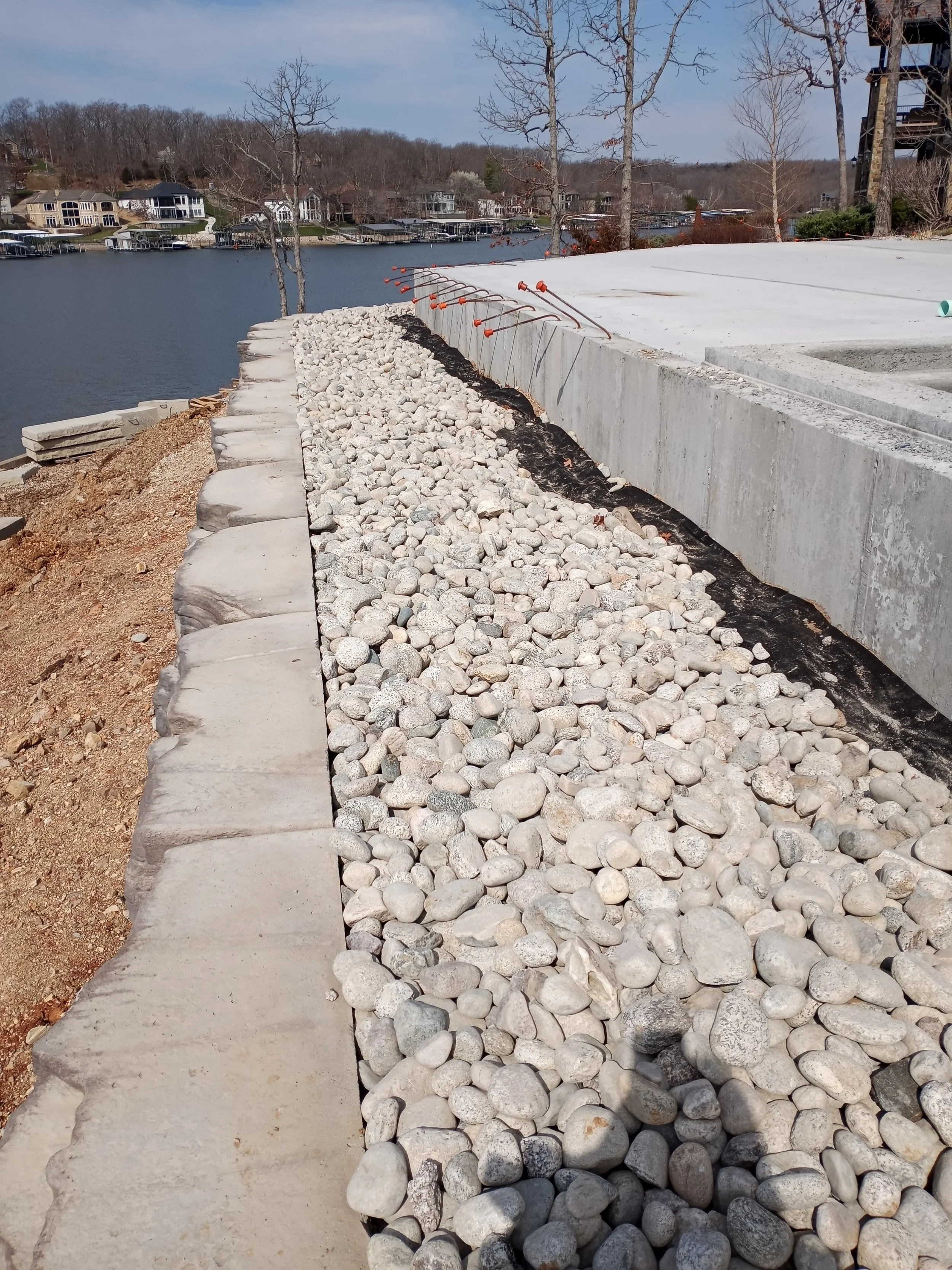 Construction site by a lake with rocks, concrete, and orange safety markers for landscaping.