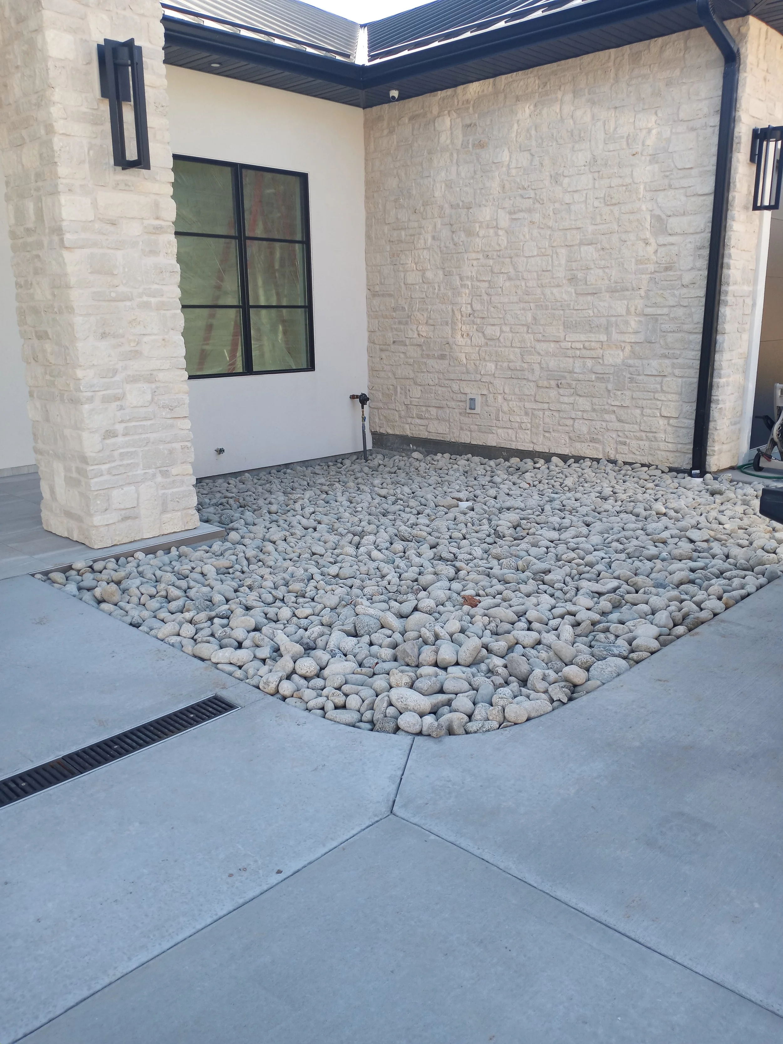 A corner of a building with a patio area, featuring gravel and stone landscaping, a beige brick wall, and a window with black framing.
