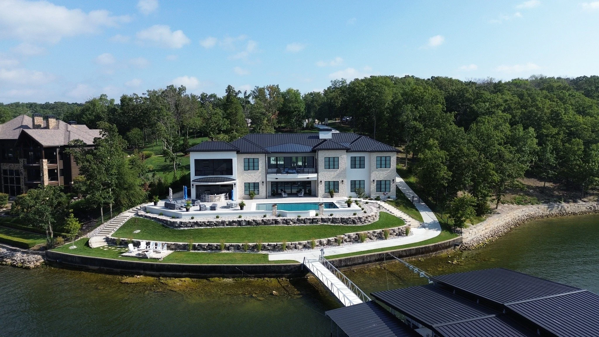 A modern house with a dark roof situated by a lake, featuring an outdoor pool, lawns, and surrounding trees.