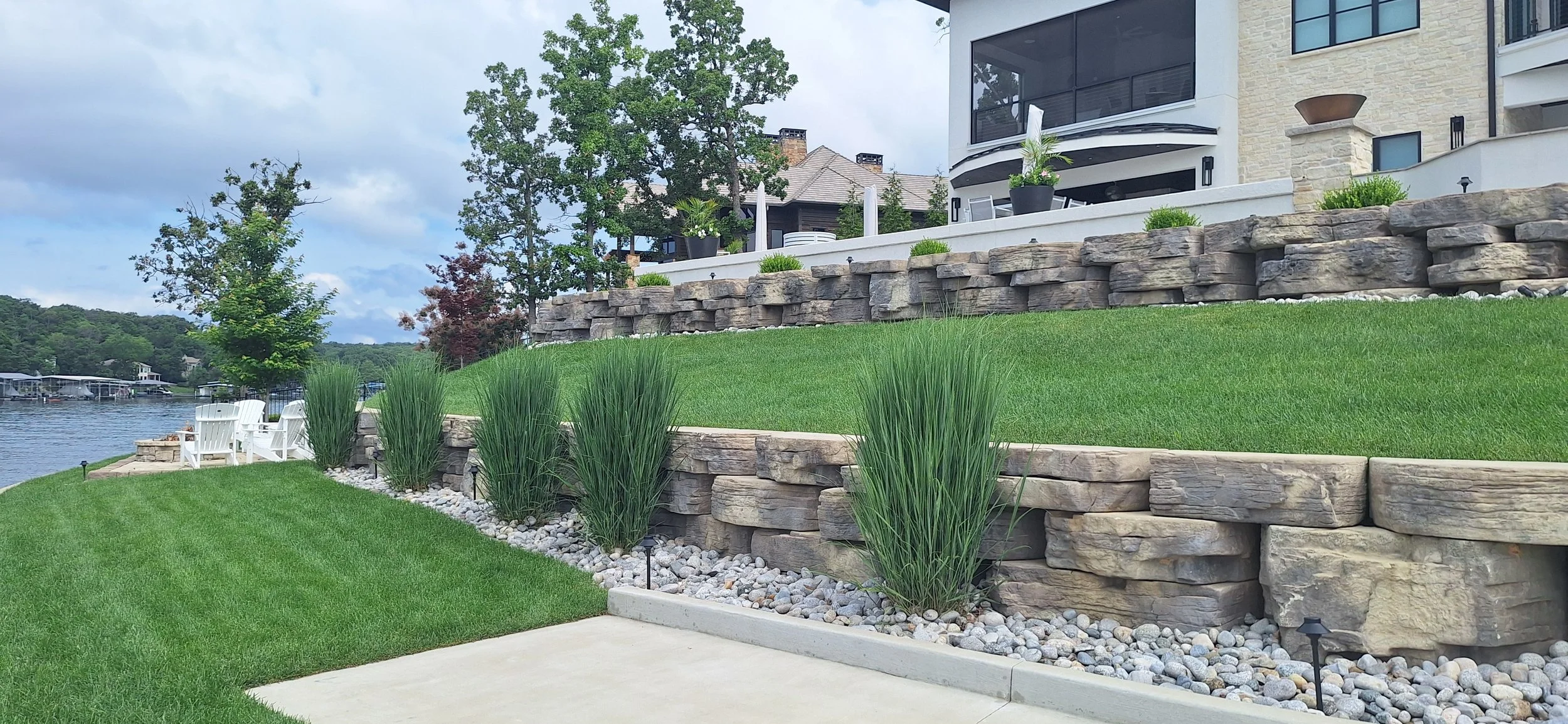 Lawn with tiered stone retaining walls, ornamental grasses, a waterfront view, and a modern house with deck and potted plants.