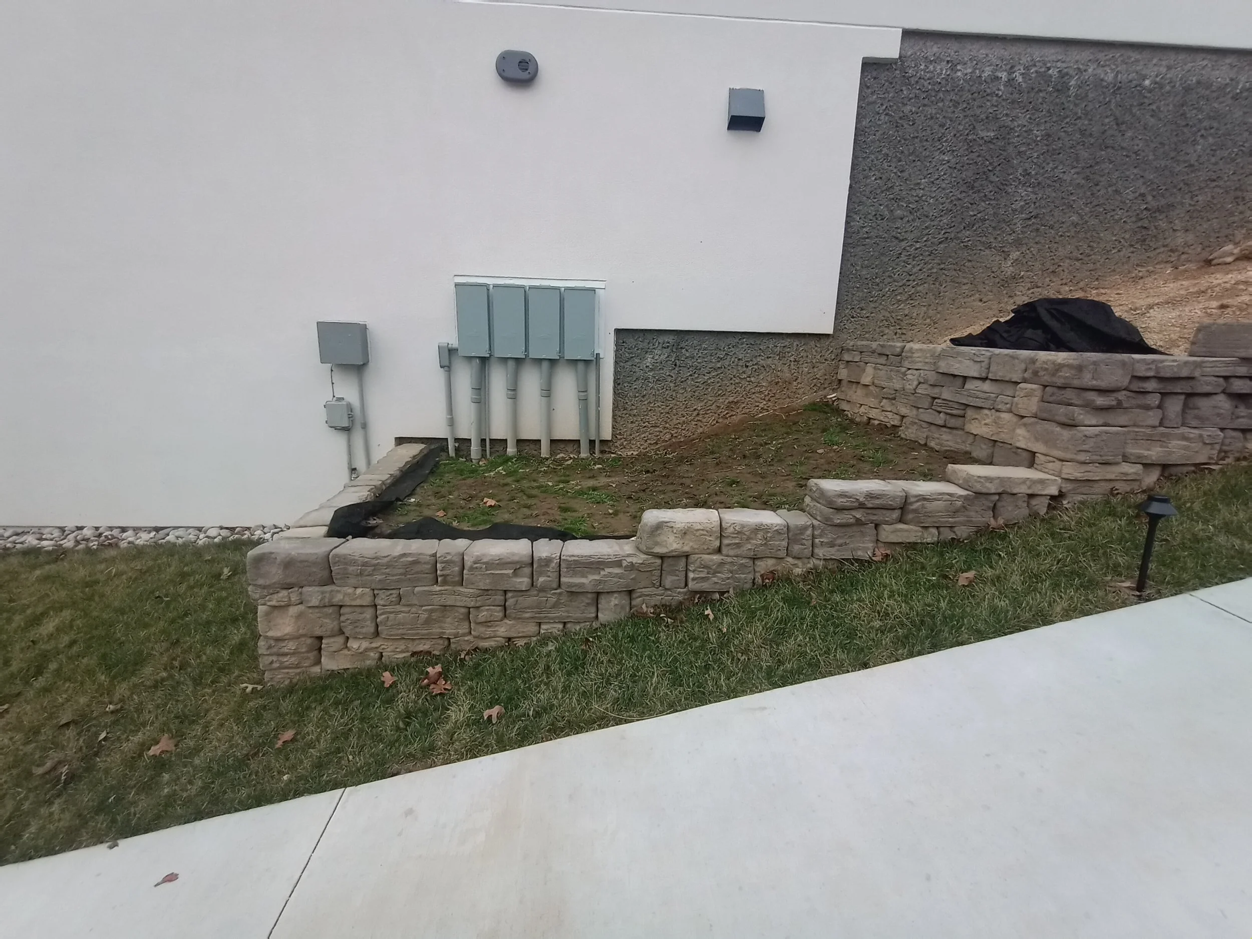 Empty yard with a partially built stone retaining wall, electrical boxes and conduit on the exterior wall of a building, and a small black landscape light on the ground.