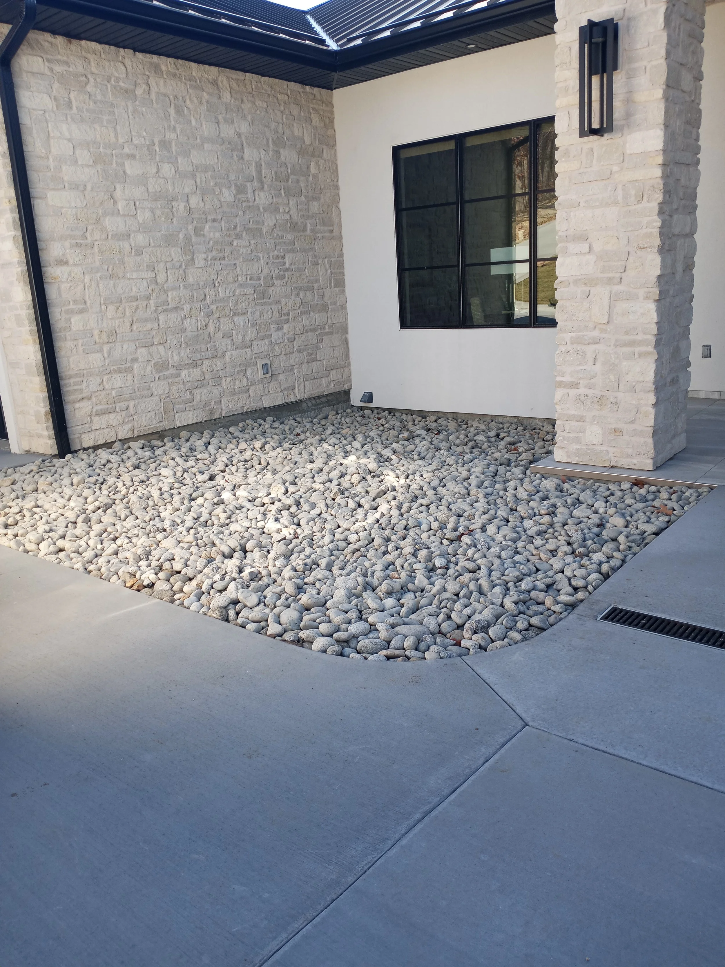 Exterior corner of a modern house with white brick and smooth stucco walls, a large black-framed window, and a garden area filled with smooth gray rocks, bordered by concrete sidewalk.