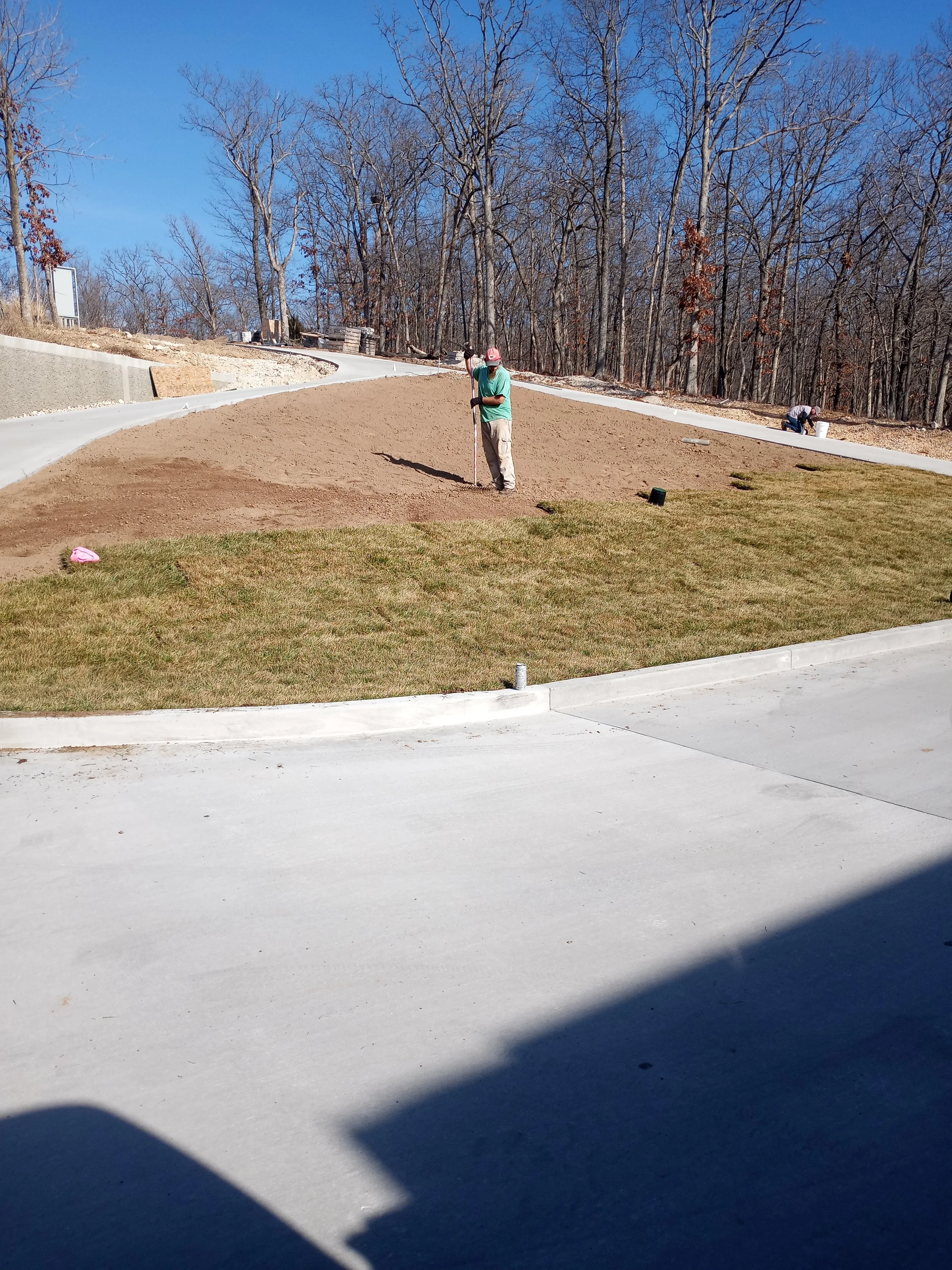 Individuals working on landscaping or gardening in a backyard, with a mix of grass and freshly tilled soil, surrounded by a wooded area with leafless trees.