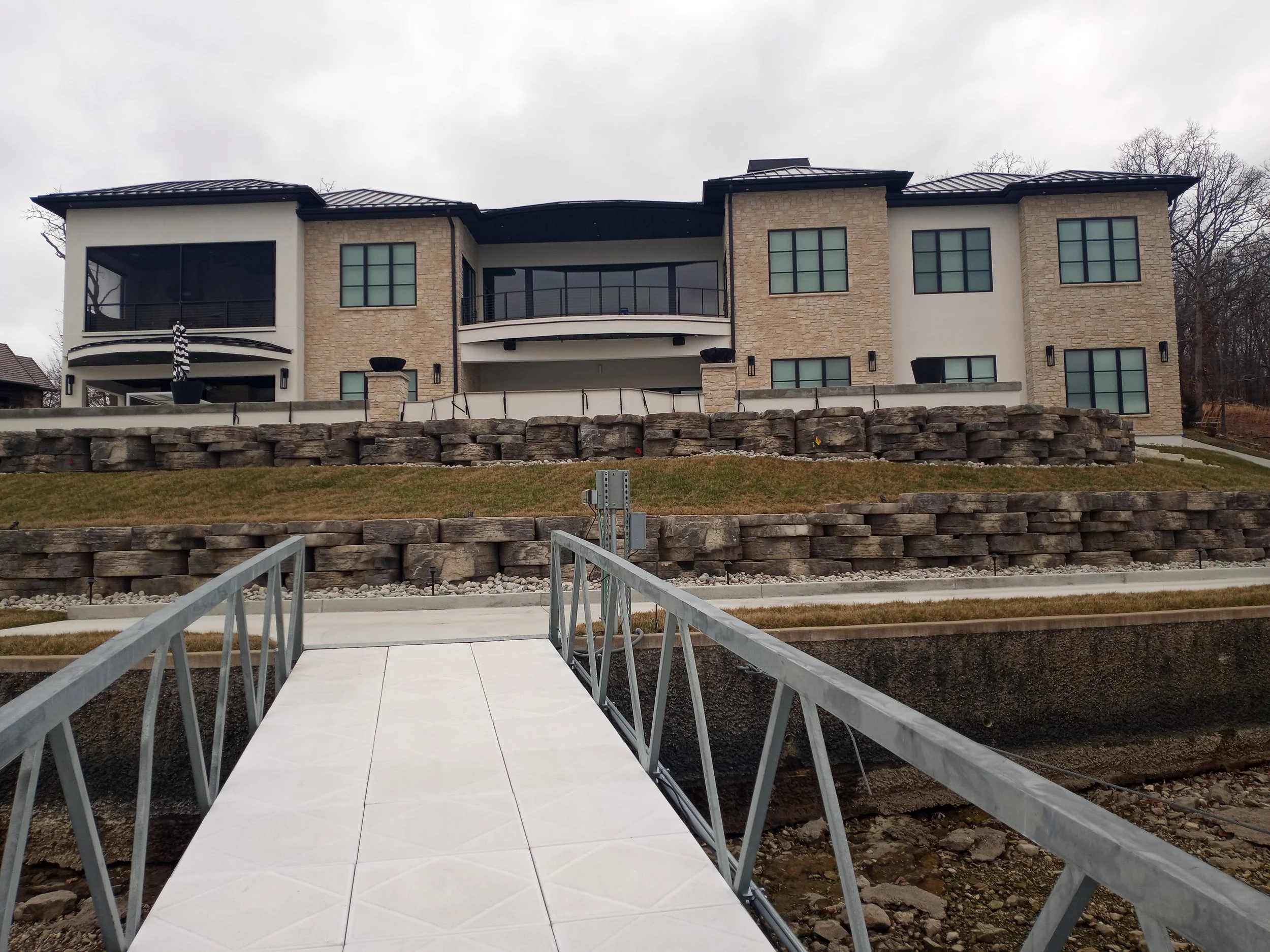 Modern multi-story residential building with large windows and balconies, situated on a hill with stone retaining walls, accessed via a small bridge.