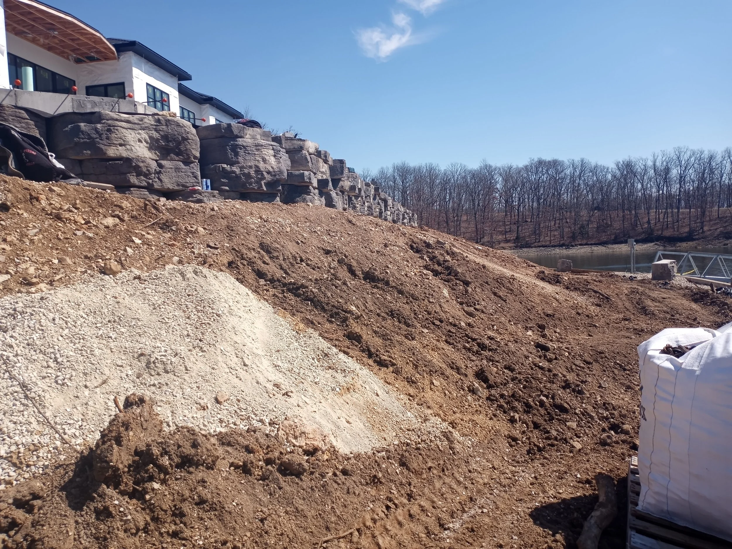 Construction site with dirt and gravel slopes, large rocks, a white house-like building at the top, trees in the background, and construction materials on the ground.