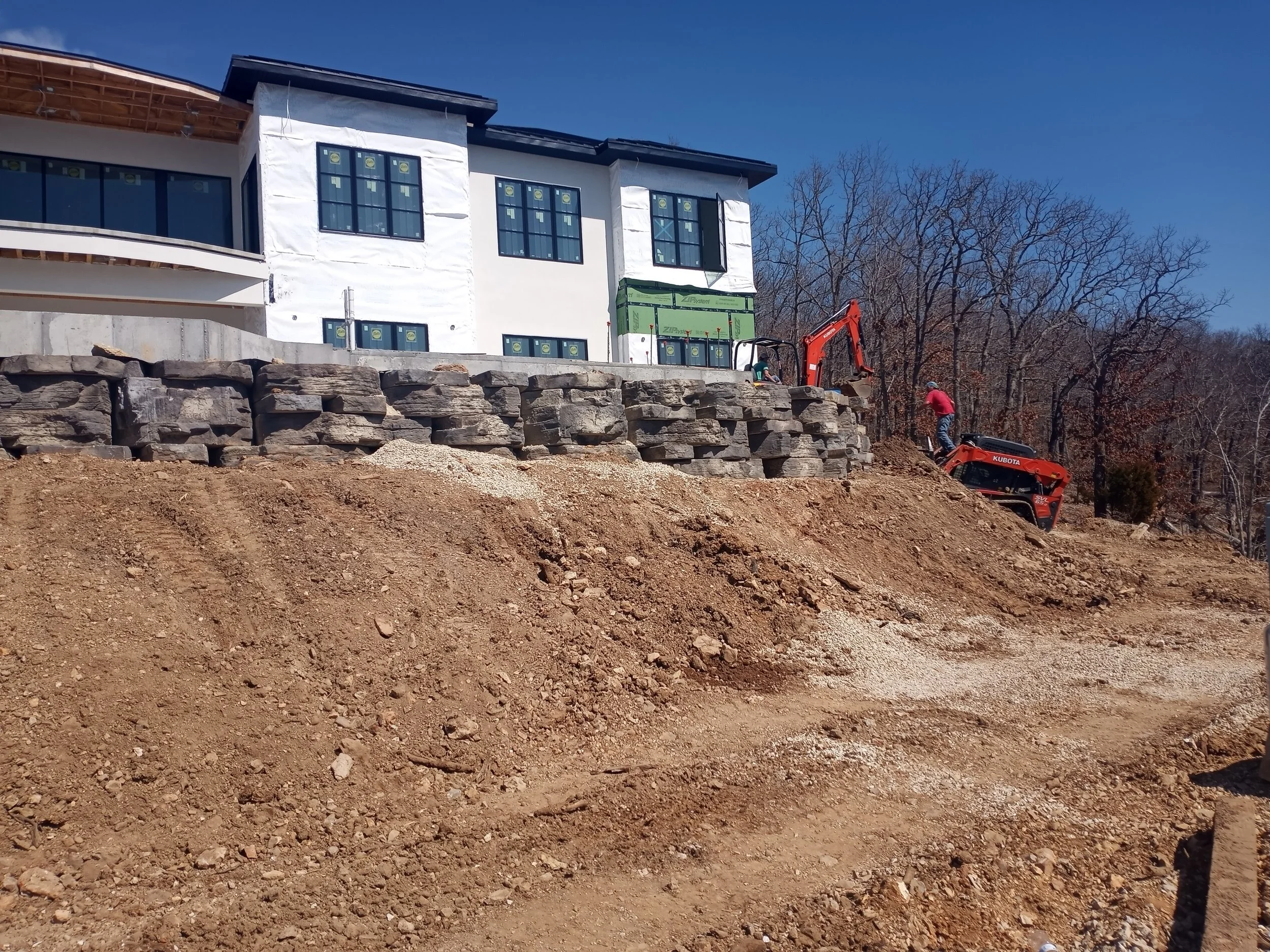 Construction site showing a house under renovation with workers and equipment on a dirt hill, with trees in the background.