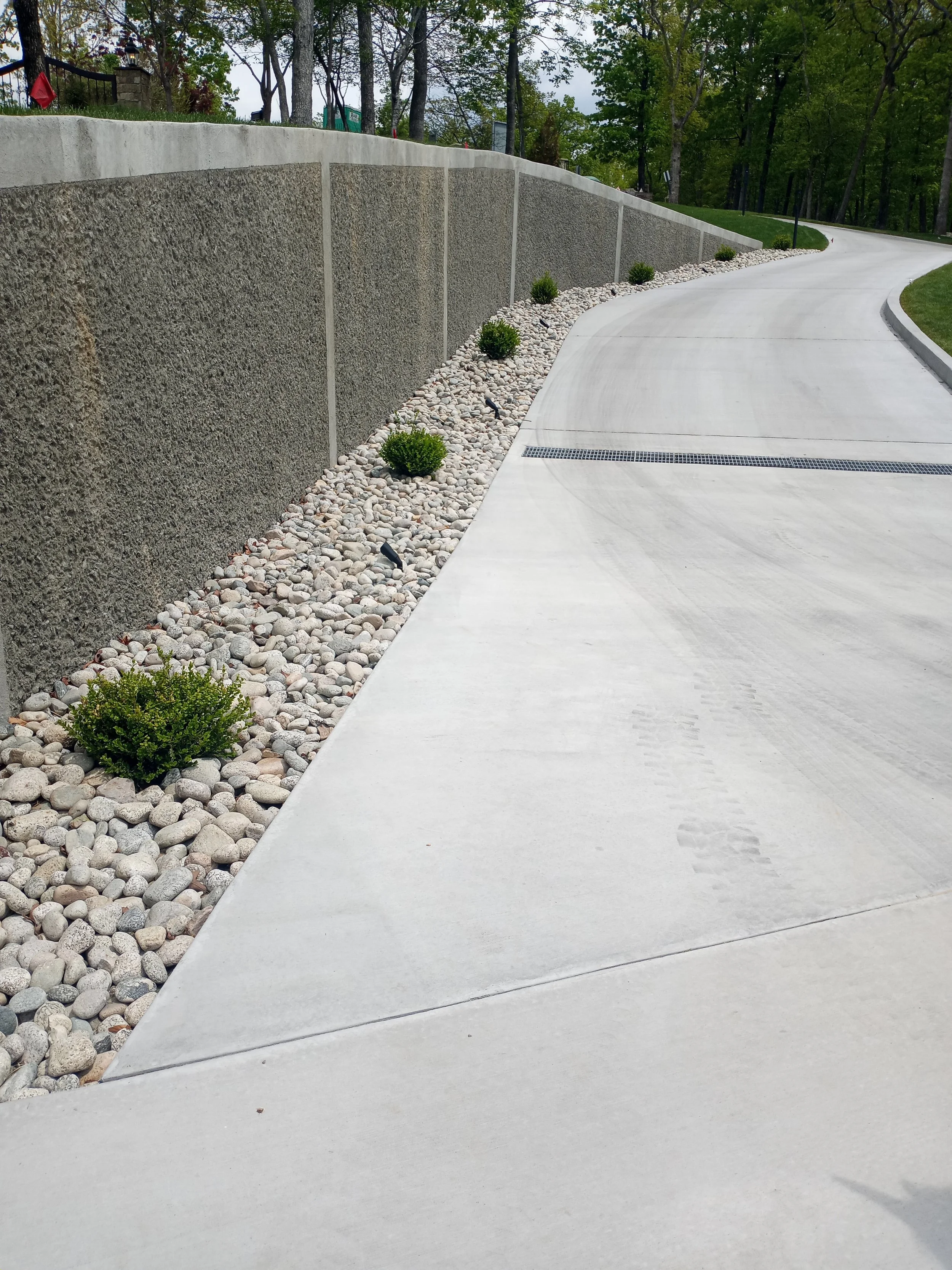 Curved driveway with gravel and small bushes along a concrete wall and grassy area with trees.