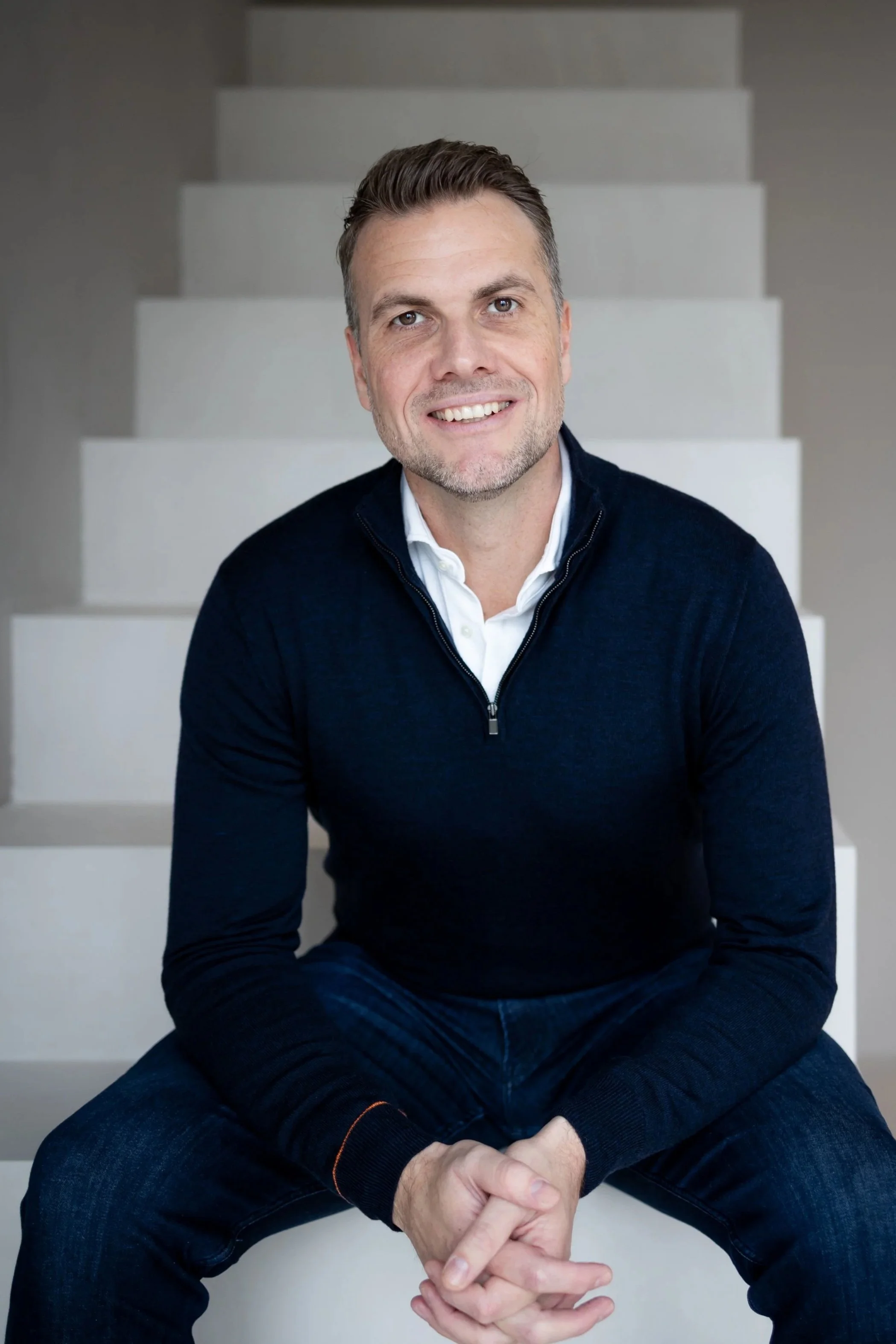 een man zit op de treden van een witte trap, lachend en gekleed in een donkerblauwe trui en een witte blouse.