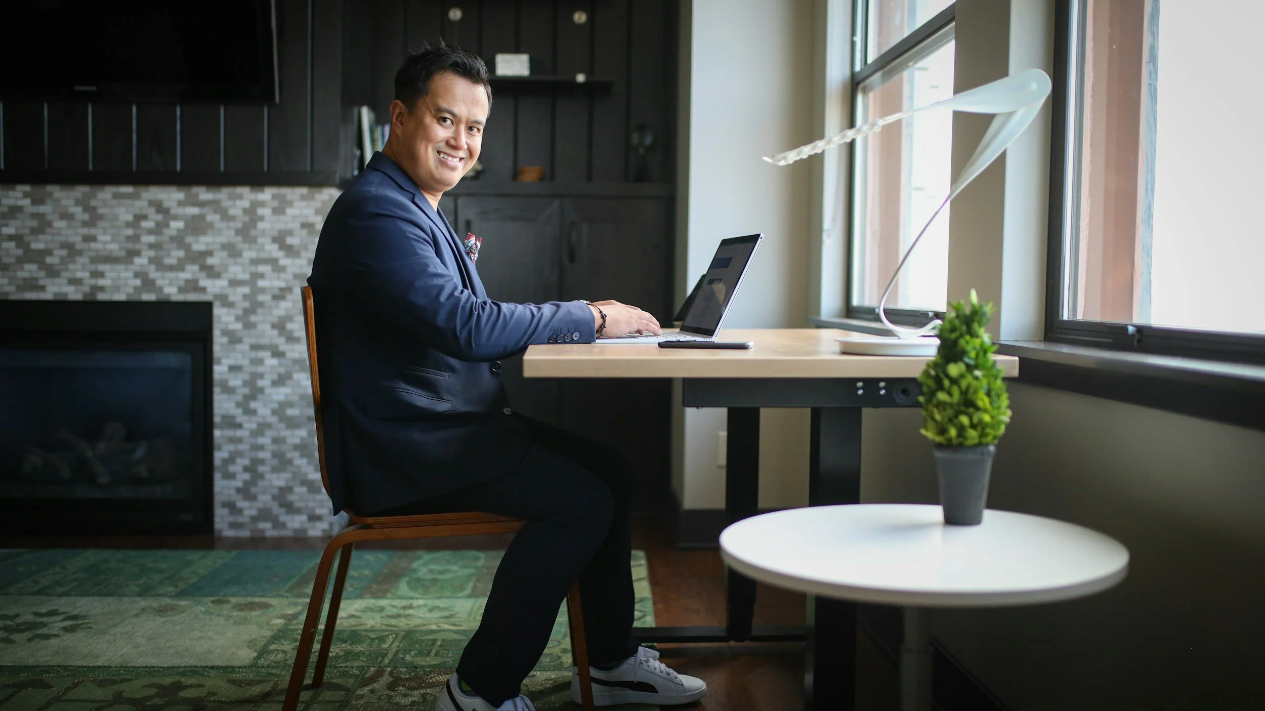 A man in a navy blazer sitting at a desk with a laptop, smiling, in a modern room with large windows and a green plant on a small white table.
