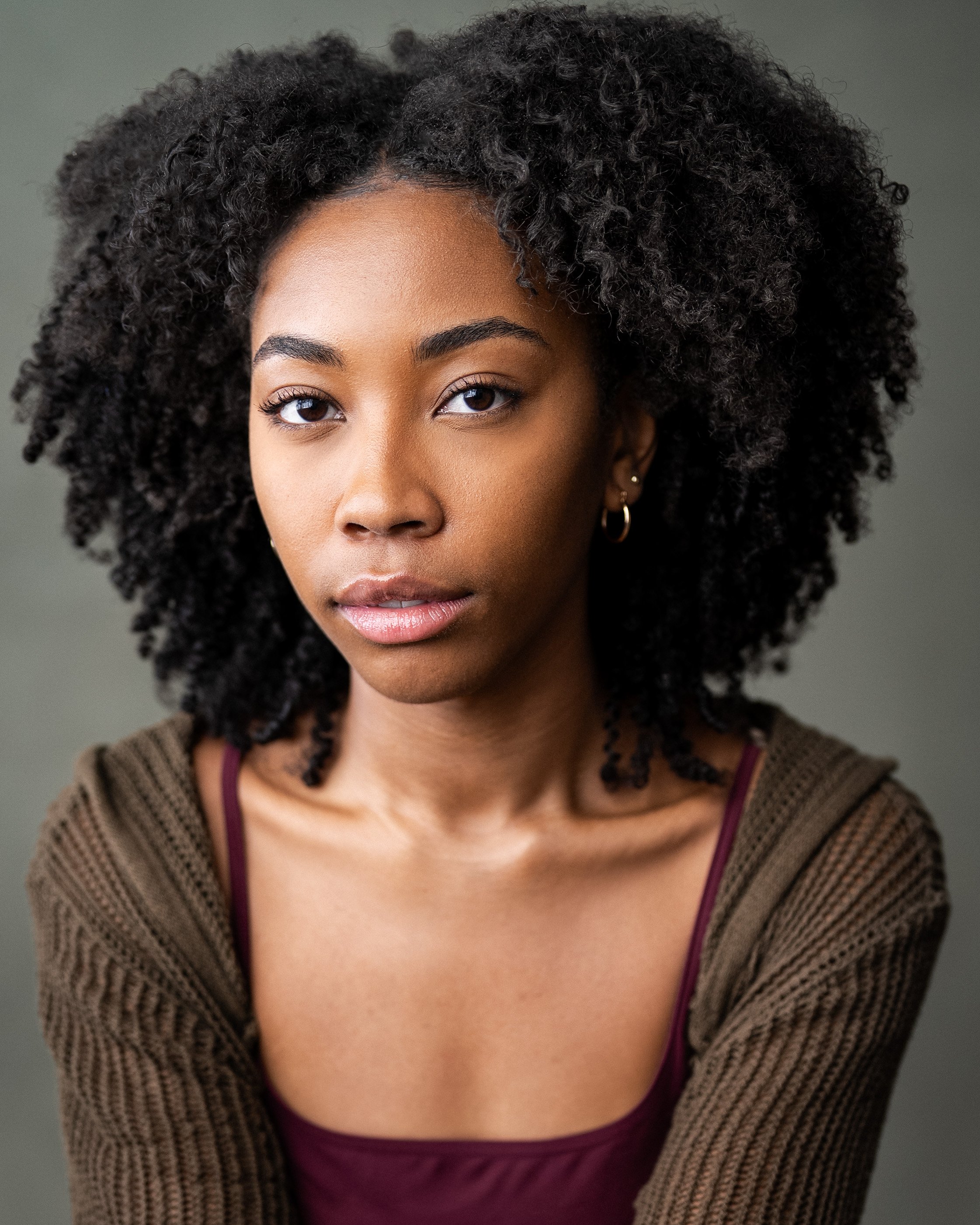 Headshot of a Nicole Stewart with curly hair, wearing hoop earrings and a green crochet sweater over a burgundy top, against a neutral background.