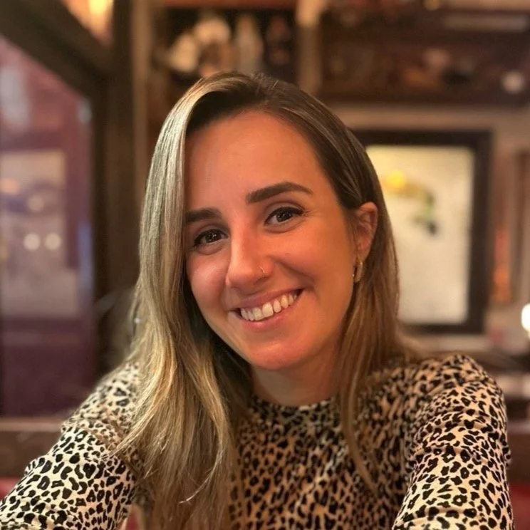 Nicole Caetano headshot. Smiling woman with long hair, wearing leopard print top, sitting indoors.
