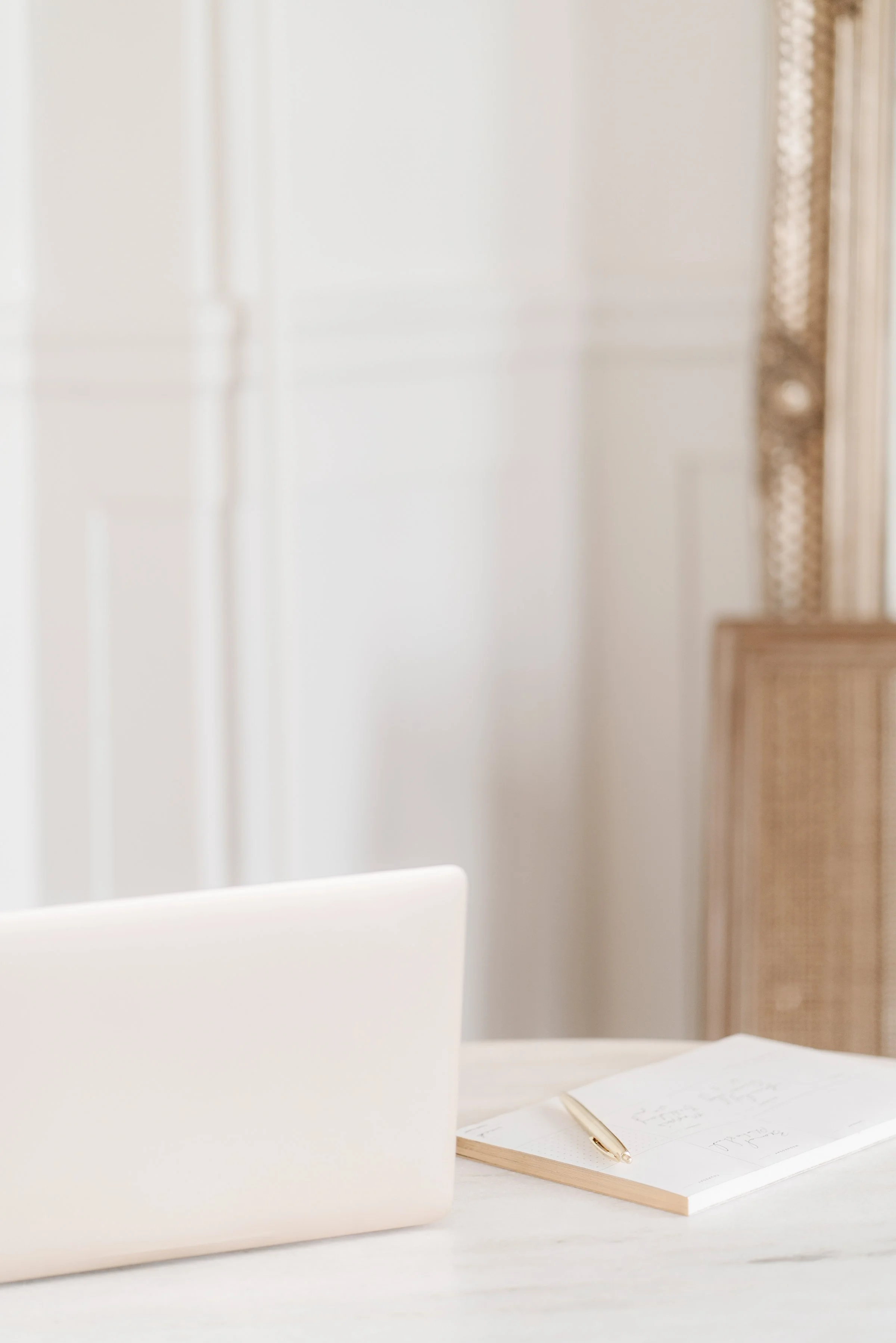 Minimalist workspace with a closed white laptop, a notebook, and a gold pen on a white table in front of a white paneled wall and a wicker cabinet.