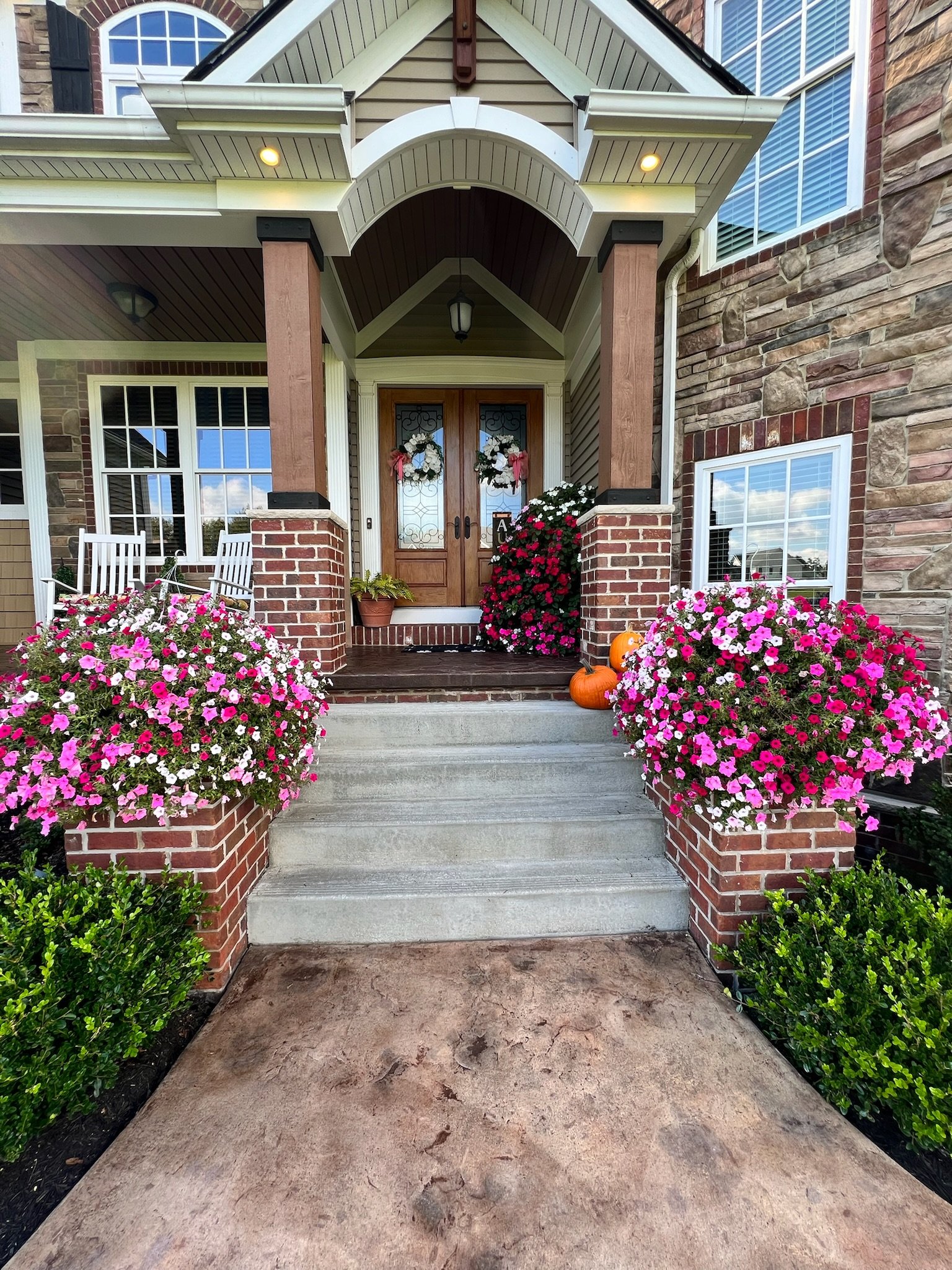 Front porch of a house with brick and stone decor, featuring steps leading to a wooden door. The porch is adorned with pink and white flowers in planters and two pumpkins. There are rocking chairs to the left and hanging lamp above the entrance.