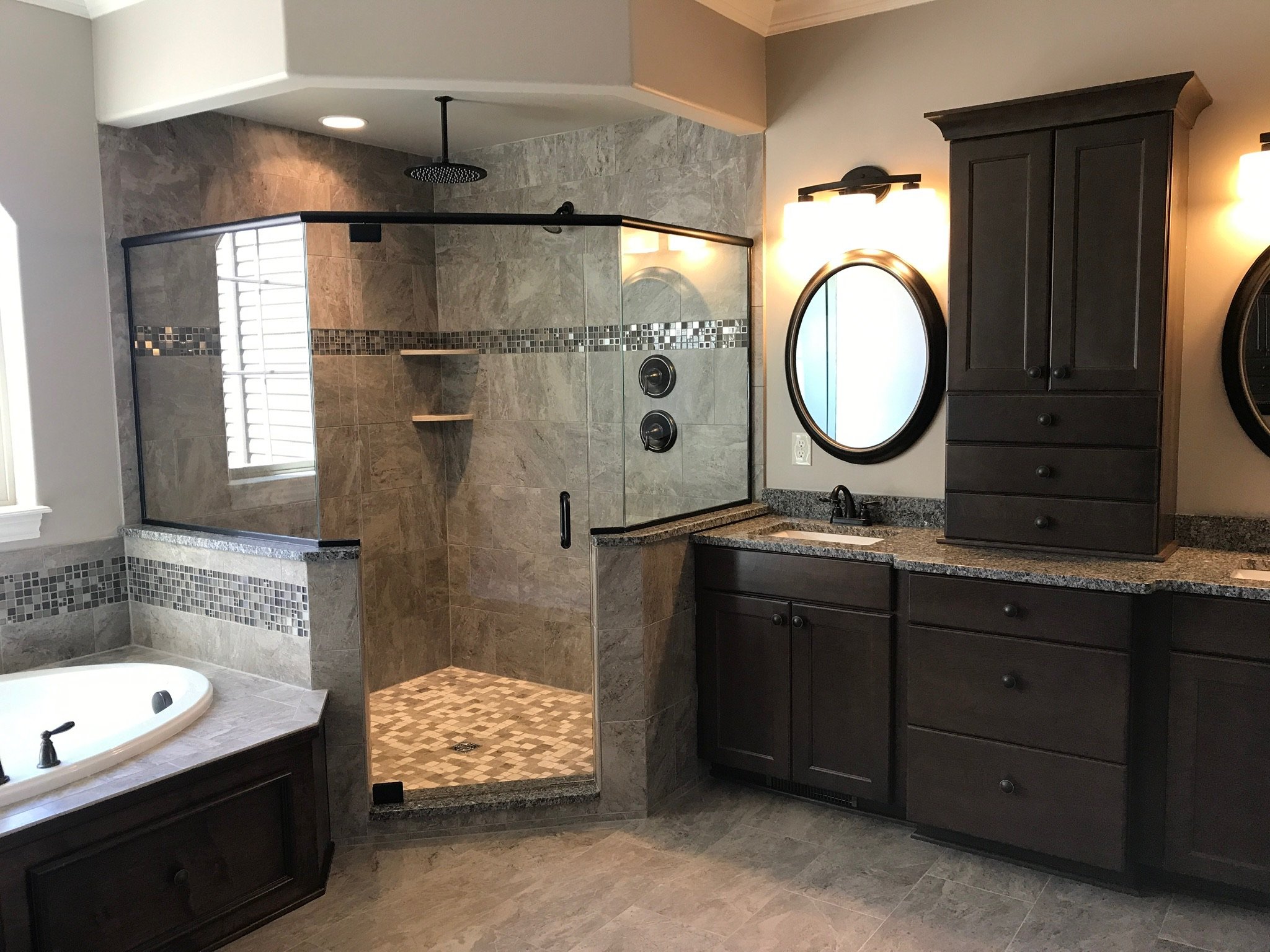 Modern bathroom with glass shower enclosure, dark wood cabinets, granite countertops, round mirrors, bathtub, and tile flooring.
