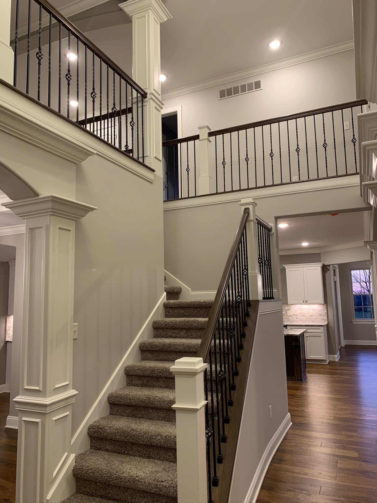 Modern interior with a carpeted staircase, elegant iron railings, and wooden flooring. The space is well-lit with recessed lighting, and the open layout reveals a kitchen area in the background.