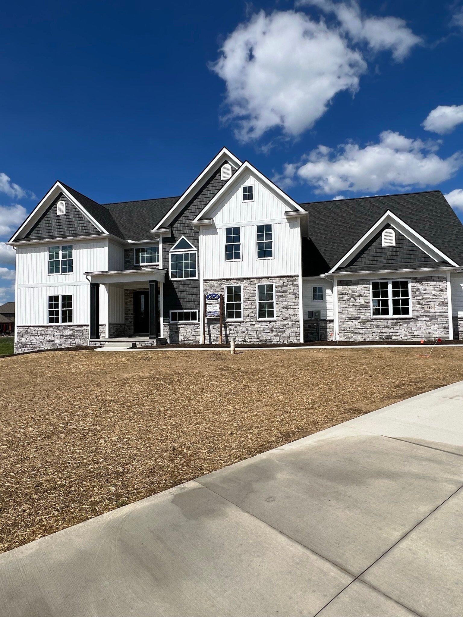 Two-story modern house with a stone facade, a black roof, and white siding, situated on a freshly landscaped yard with a concrete driveway under a blue sky with clouds.