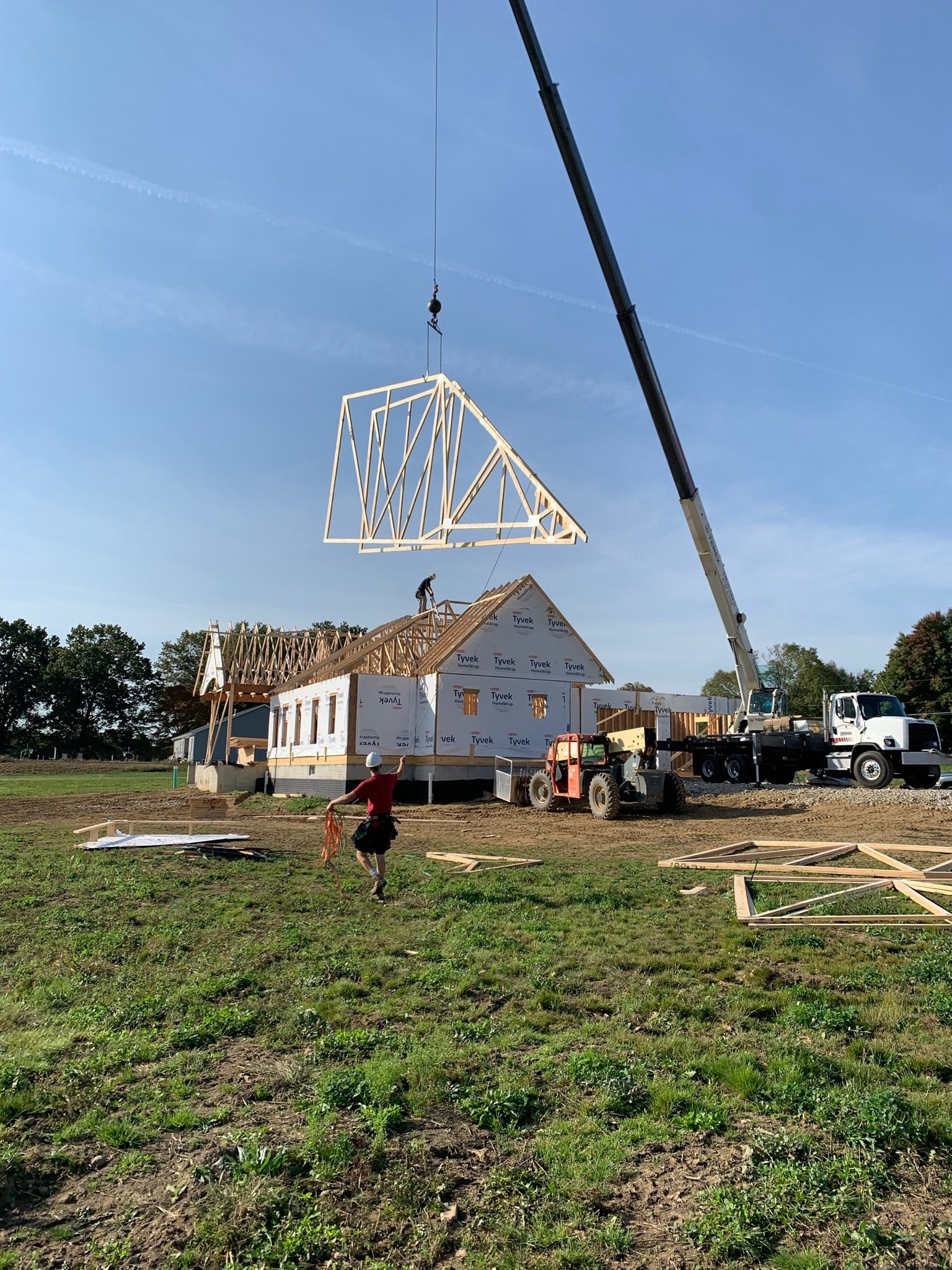 Construction site with a crane lifting a roof truss onto a partially built house. A worker is on the roof and another person is walking on the ground, carrying materials. The house has visible insulation and building materials around.