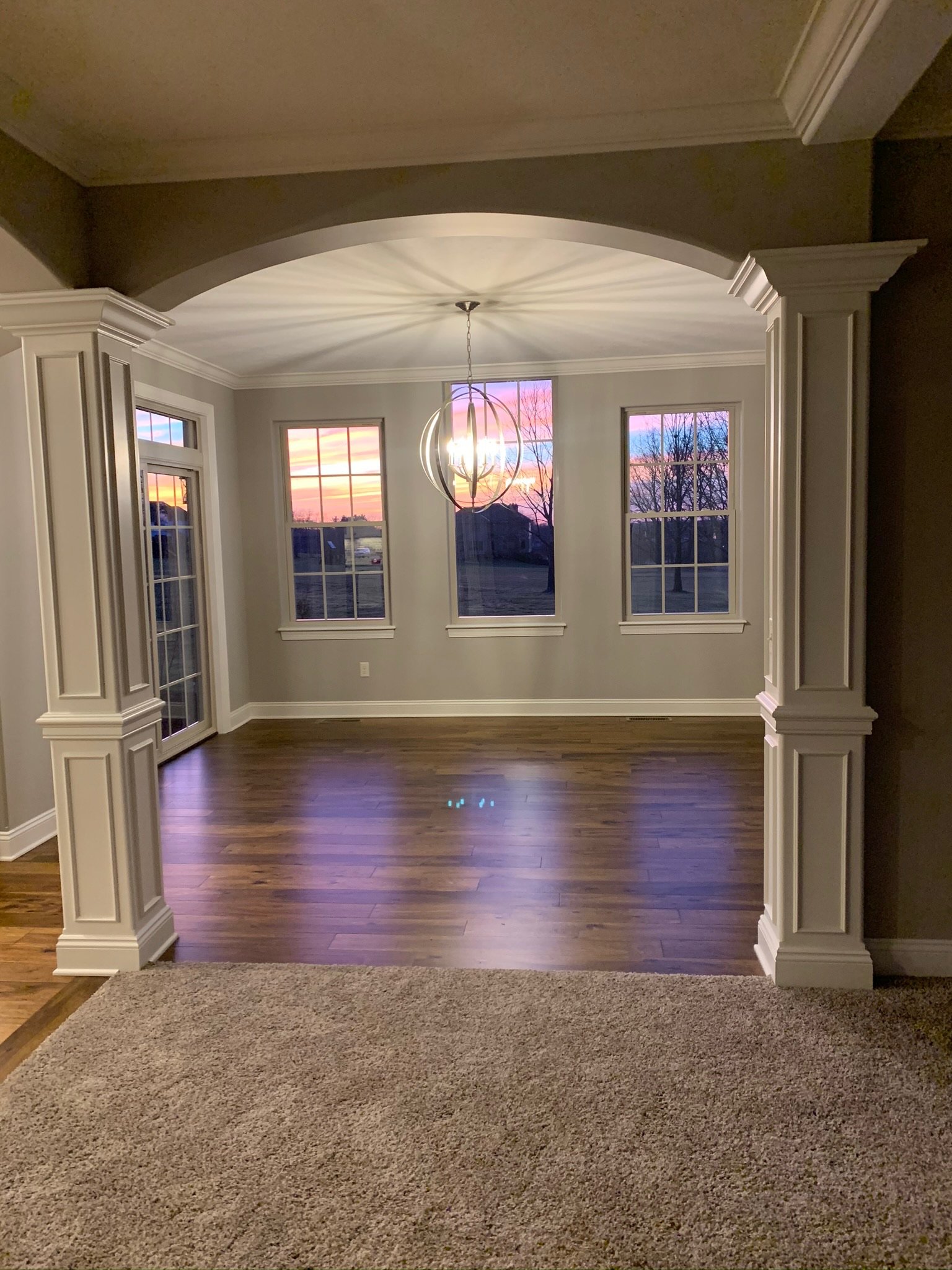 Interior view of an empty room with hardwood flooring, archway, and a modern chandelier. The room features large windows showing a colorful sunset. Carpeted area is visible in the foreground.
