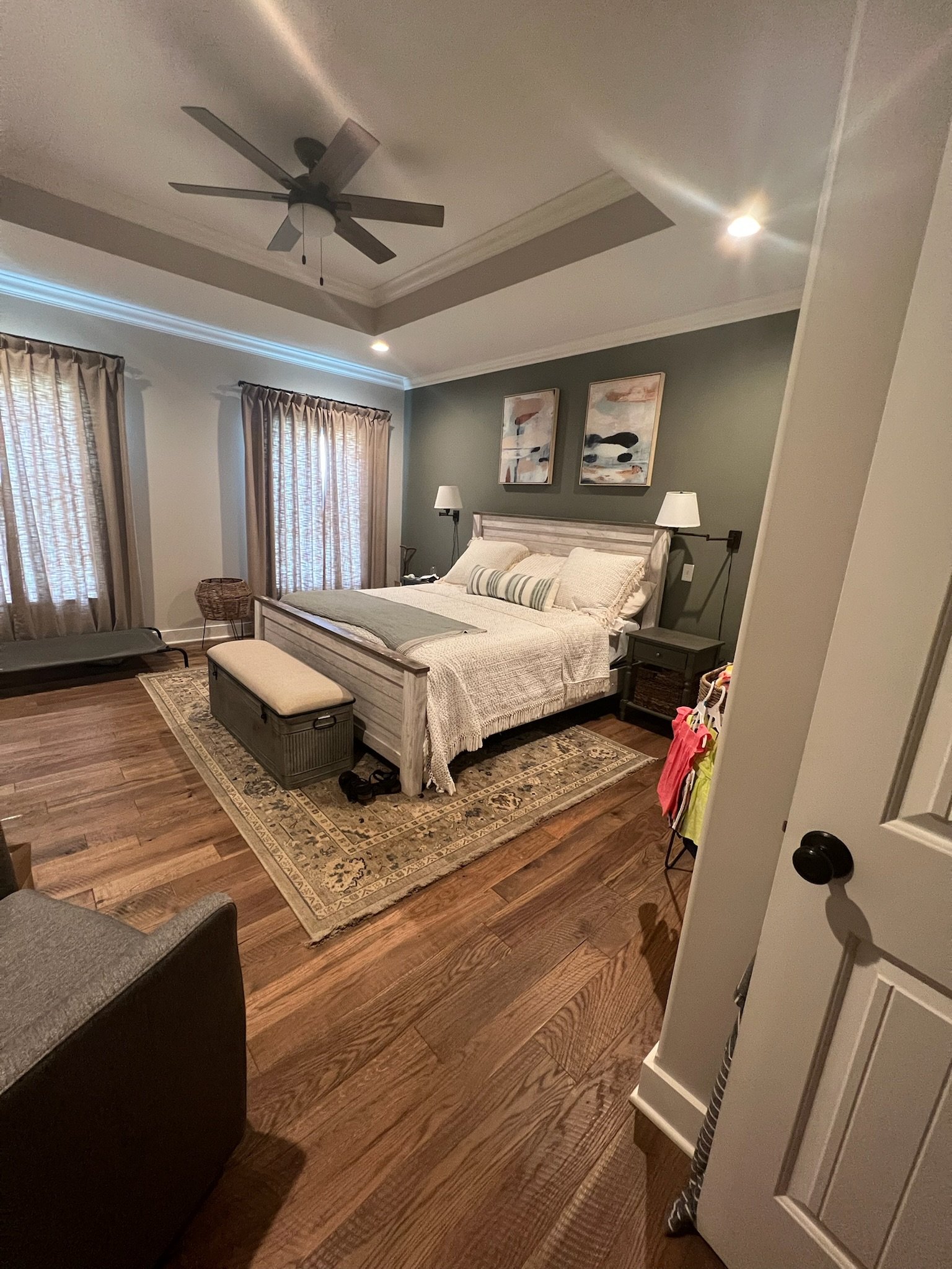 Bedroom with light wood flooring, green accent wall, and ceiling fan, featuring a bed with white bedding, two paintings, windows with curtains, a bench, and a rug.