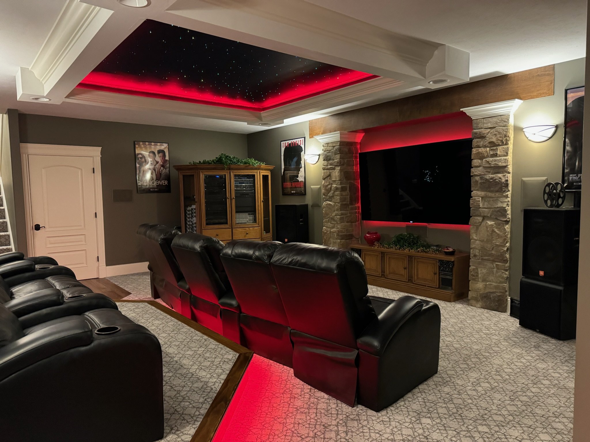 Home theater room with leather recliners, large TV, ambient red lighting, starry ceiling, and movie posters on the walls.