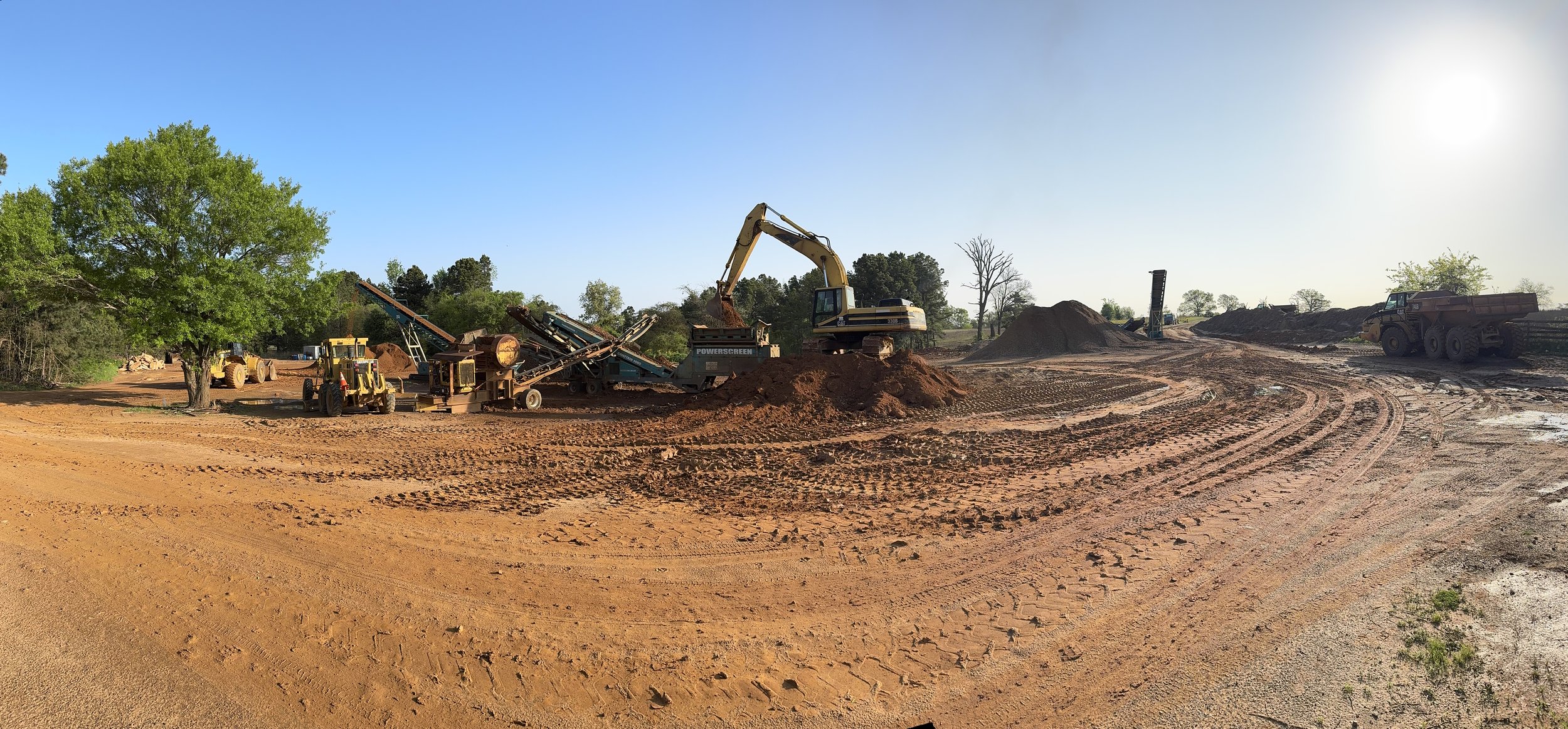 Construction site with heavy machinery, including excavators and loaders, on a dirt landscape with piled earth and trees in the background.