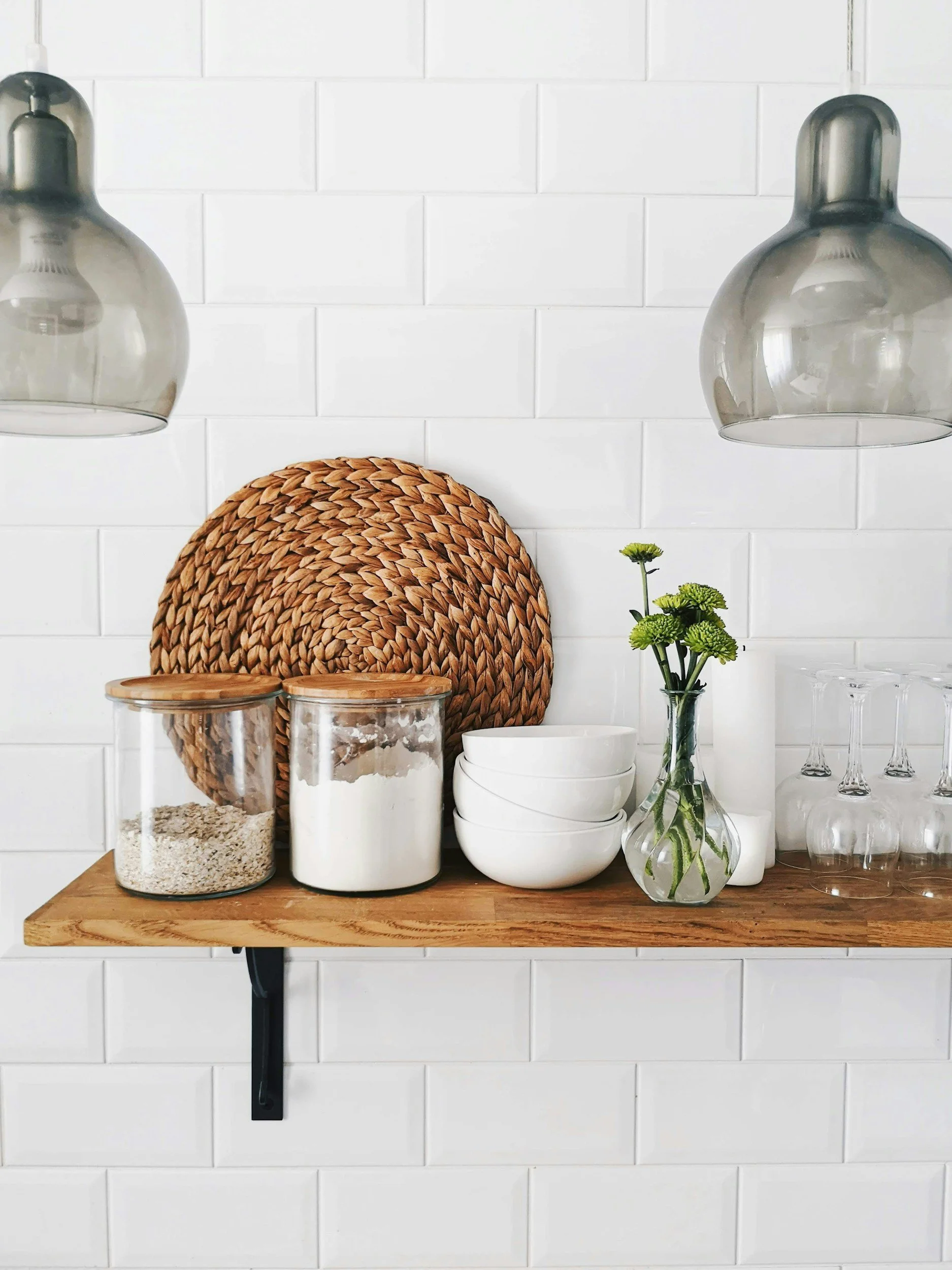 A wooden wall shelf with containers of oats and flour, stacked white bowls, a small vase with green flowers, and empty wine glasses, against a white brick wall.