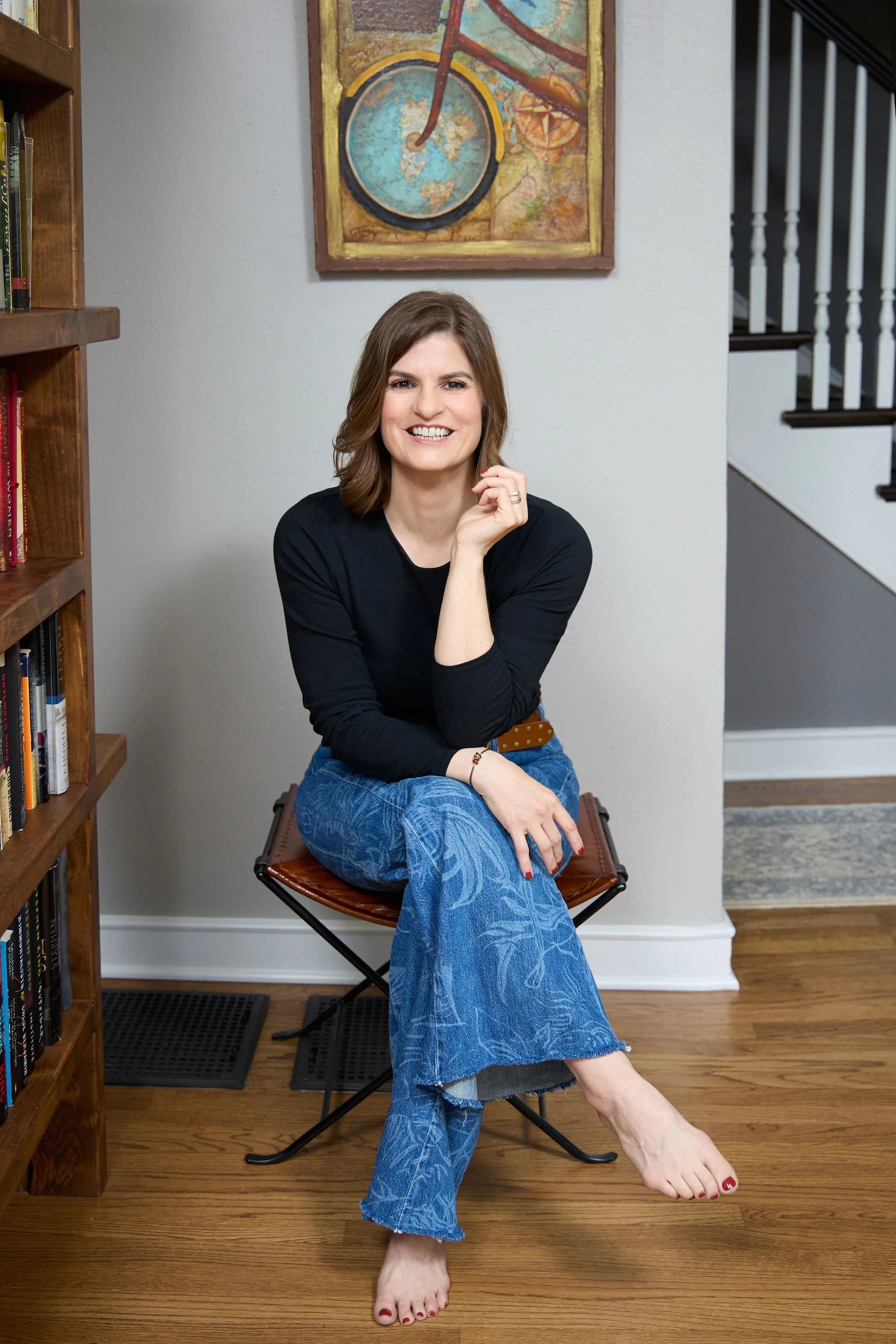 A smiling woman with shoulder-length brown hair, wearing a black long-sleeve top and blue patterned jeans, sitting barefoot on a brown leather chair in a room with a wooden floor. There are bookshelves on her left and a colorful framed map or artwork on the wall behind her. She is sitting with one leg crossed over the other and resting her chin on her hand.