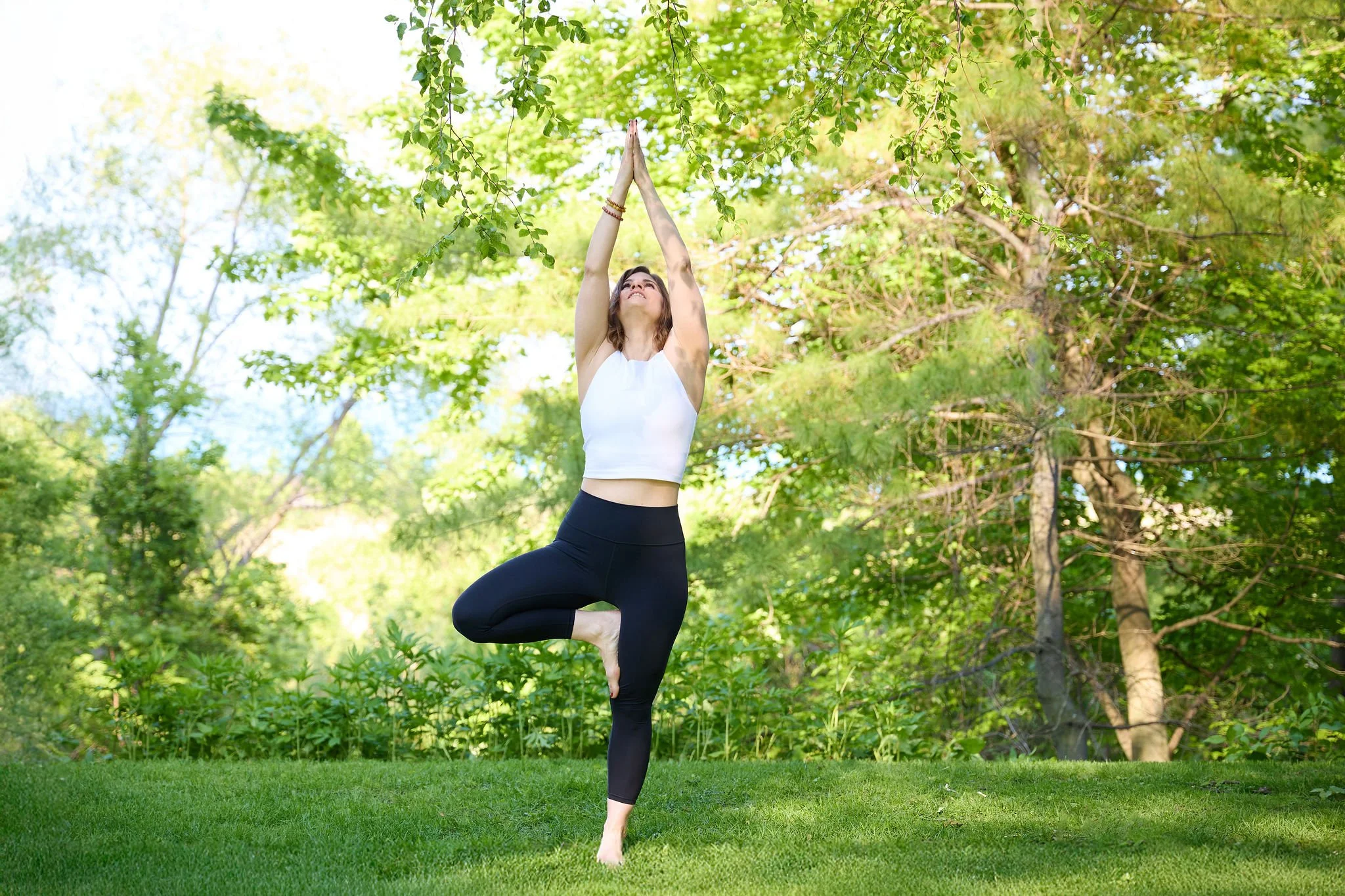 Woman practicing yoga outdoors in a tree pose on a green lawn surrounded by lush trees and foliage.