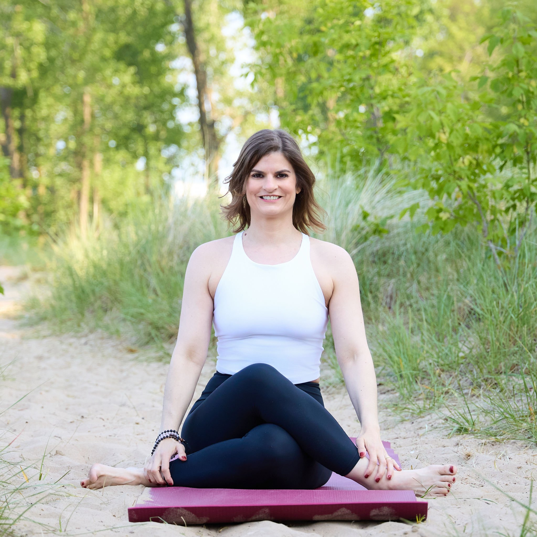 A woman with short brown hair and a white tank top is sitting cross-legged on a yoga mat in a sandy outdoor area surrounded by green trees and grass.