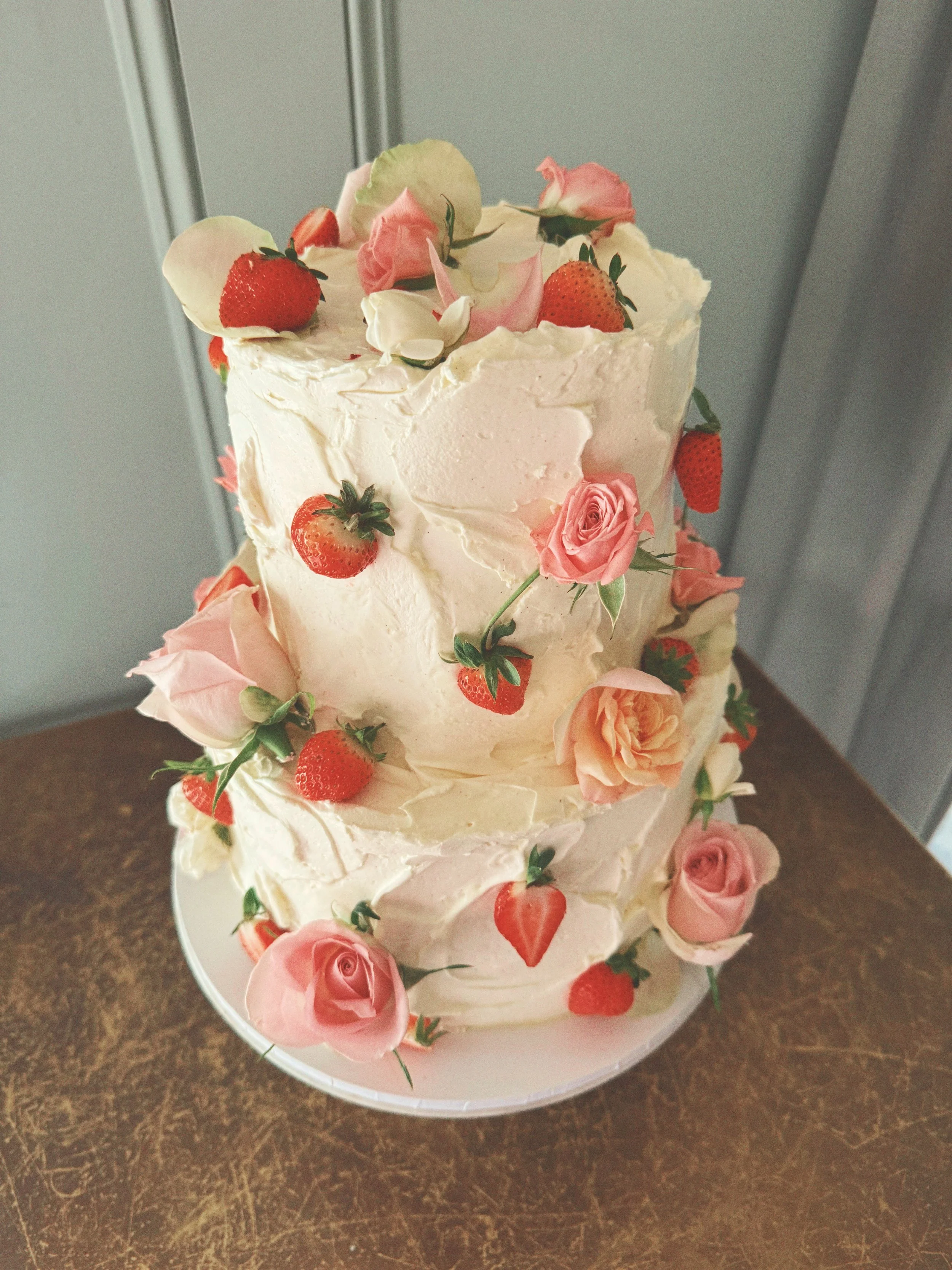 A three-tiered wedding cake decorated with fresh strawberries, pink roses, and white and pink flower petals.