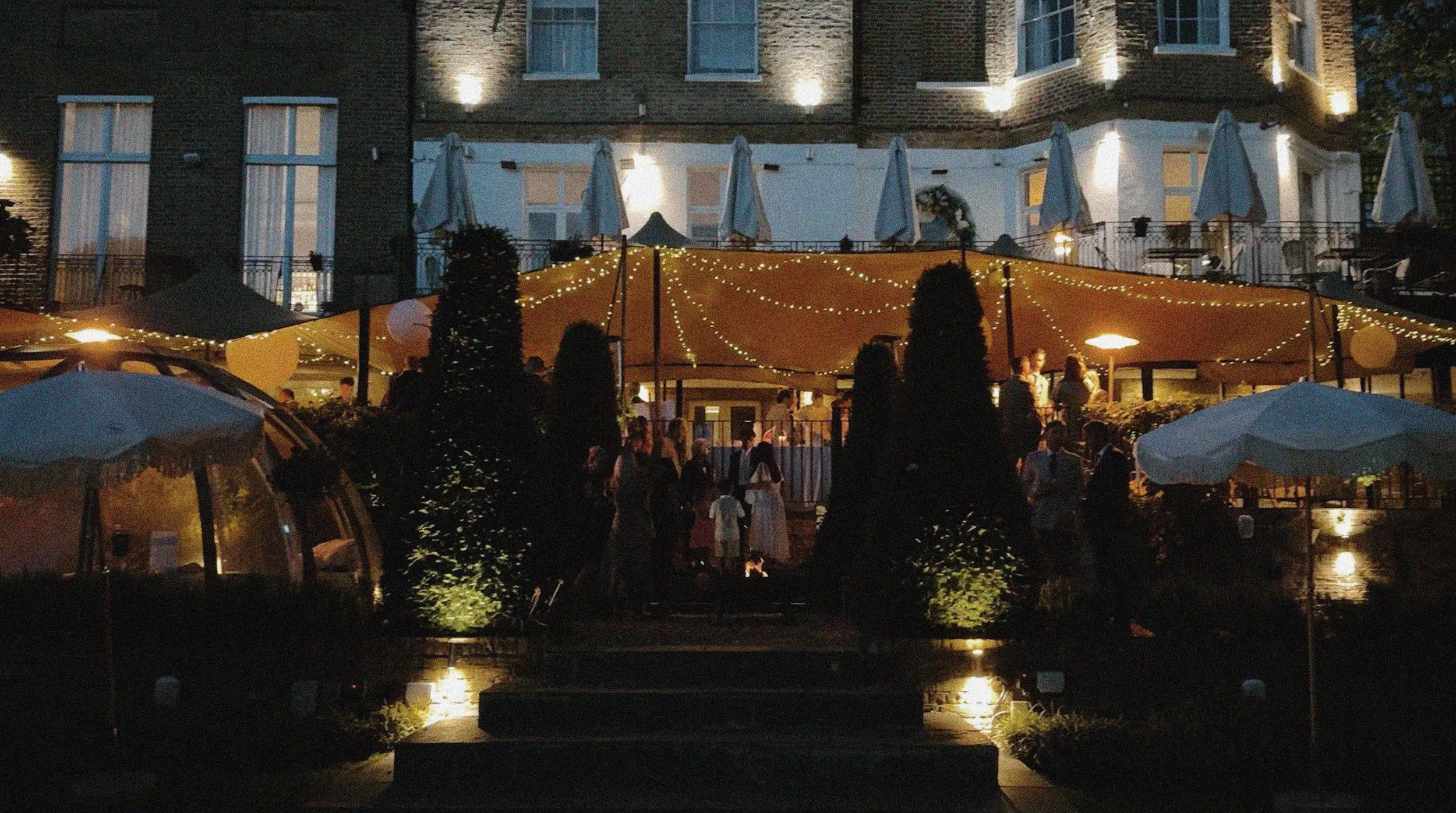 Night scene of an outdoor gathering at the Bingham Riverhouse in Richmond with string lights and umbrellas, people are mingling near the entrance of a building with lit windows.