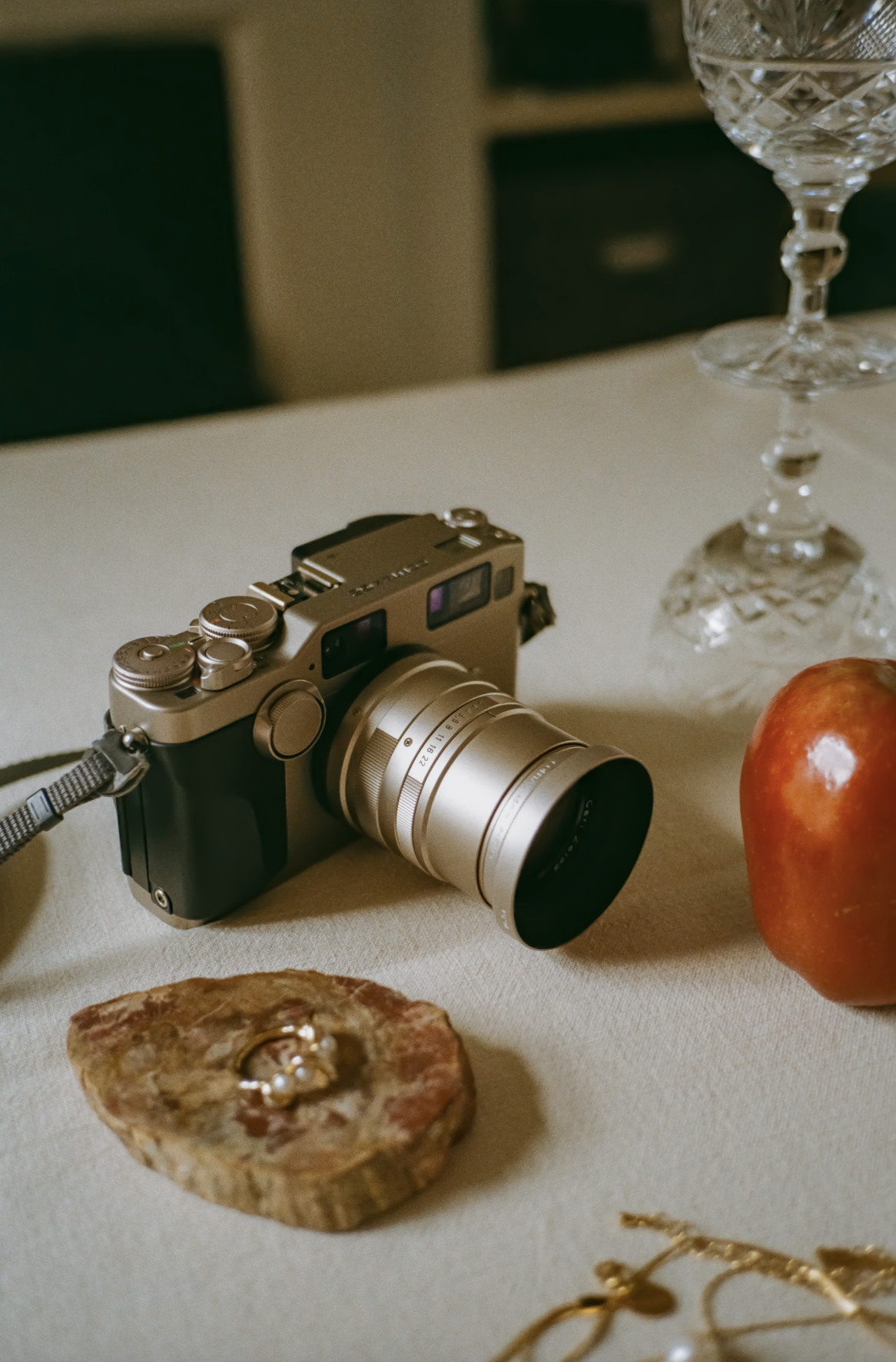 A still life scene with a camera, an apple, a piece of jewelry, and a wine glass on a table.