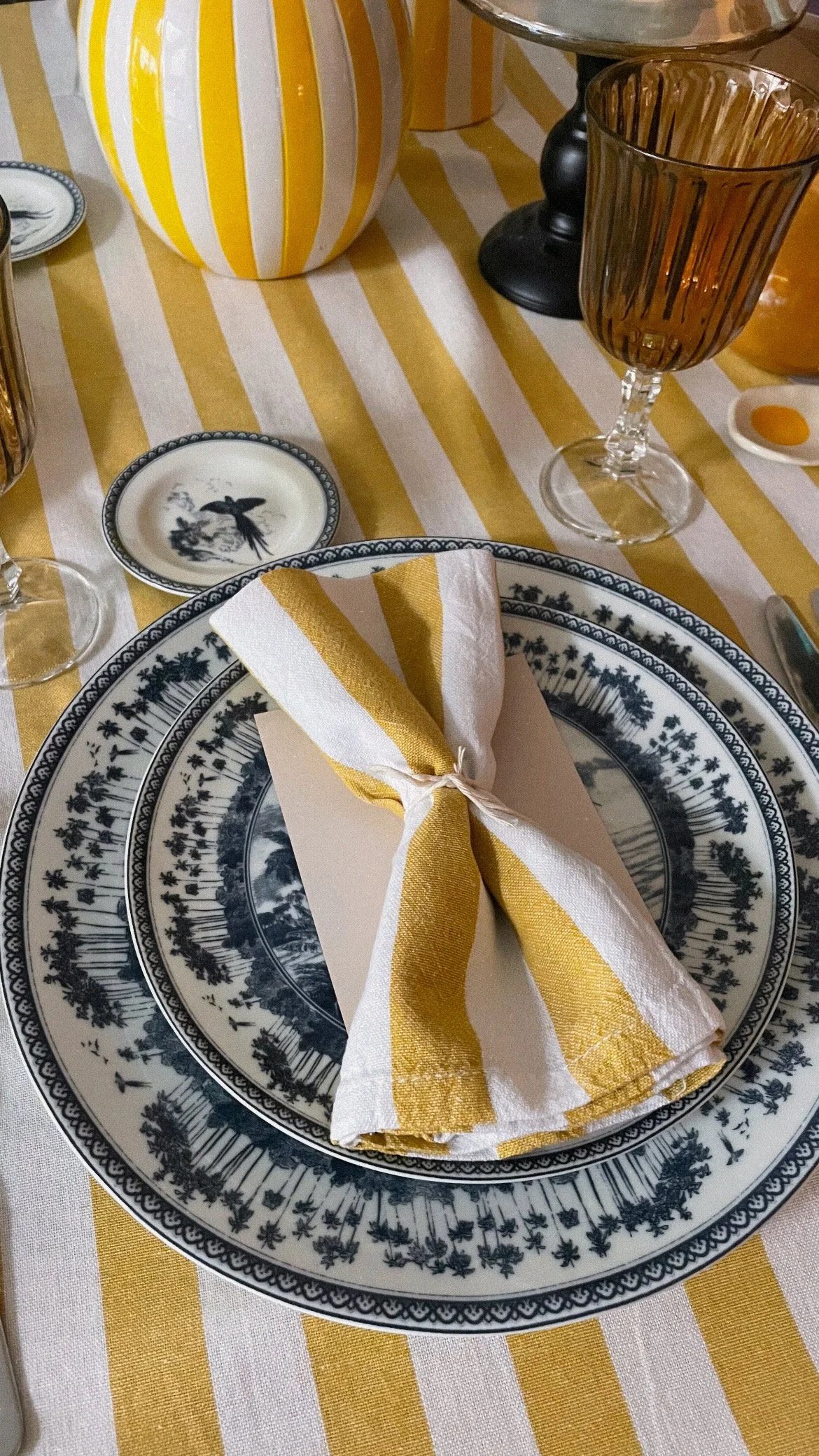 Set dining table with navy and white patterned plates, a yellow and white striped napkin tied around a card, amber-colored glasses, and a yellow and white striped ceramic vase on a yellow and white striped tablecloth.