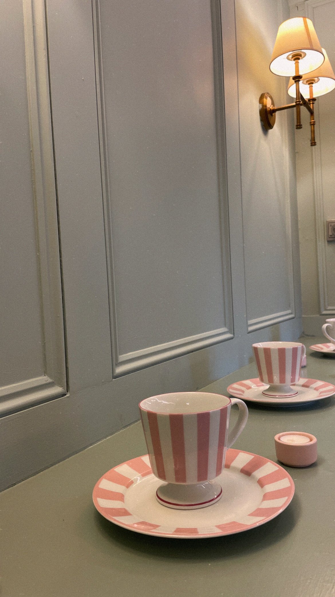 Pink striped teacups from H&M Home with matching saucers on a gray countertop, with a gray paneled wall and a wall-mounted lamp in the background.