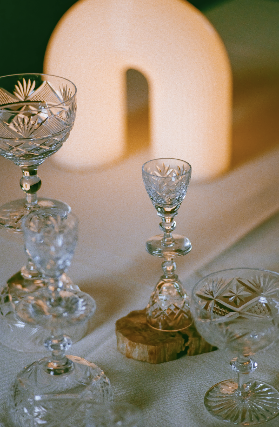 Elegant crystal glasses with intricate cut designs, displayed on a beige tablecloth, with a soft light illuminating the scene and a large illuminated letter 'O' in the background.