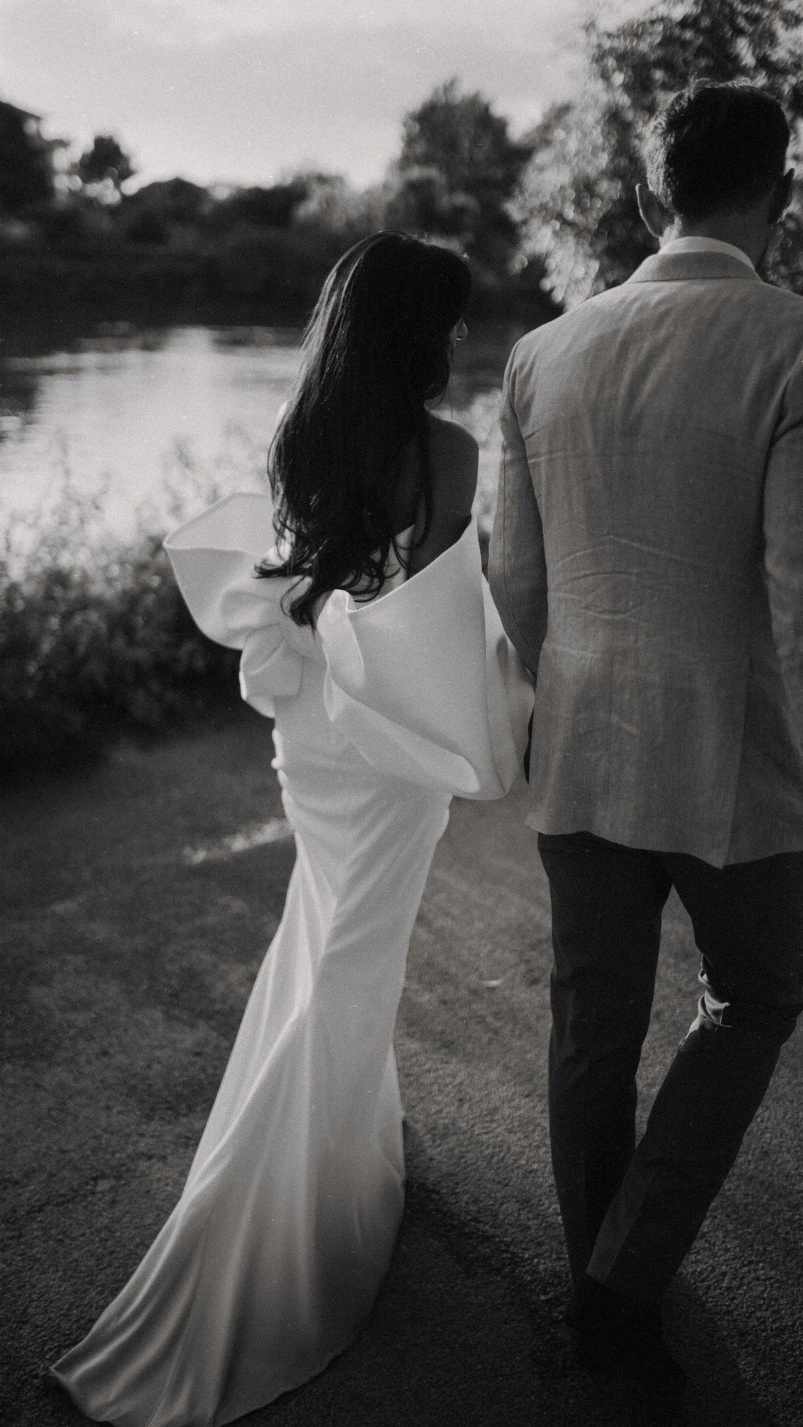 A black-and-white photo of a couple walking near a water body, with the woman wearing an elegant white gown and the man in a suit.