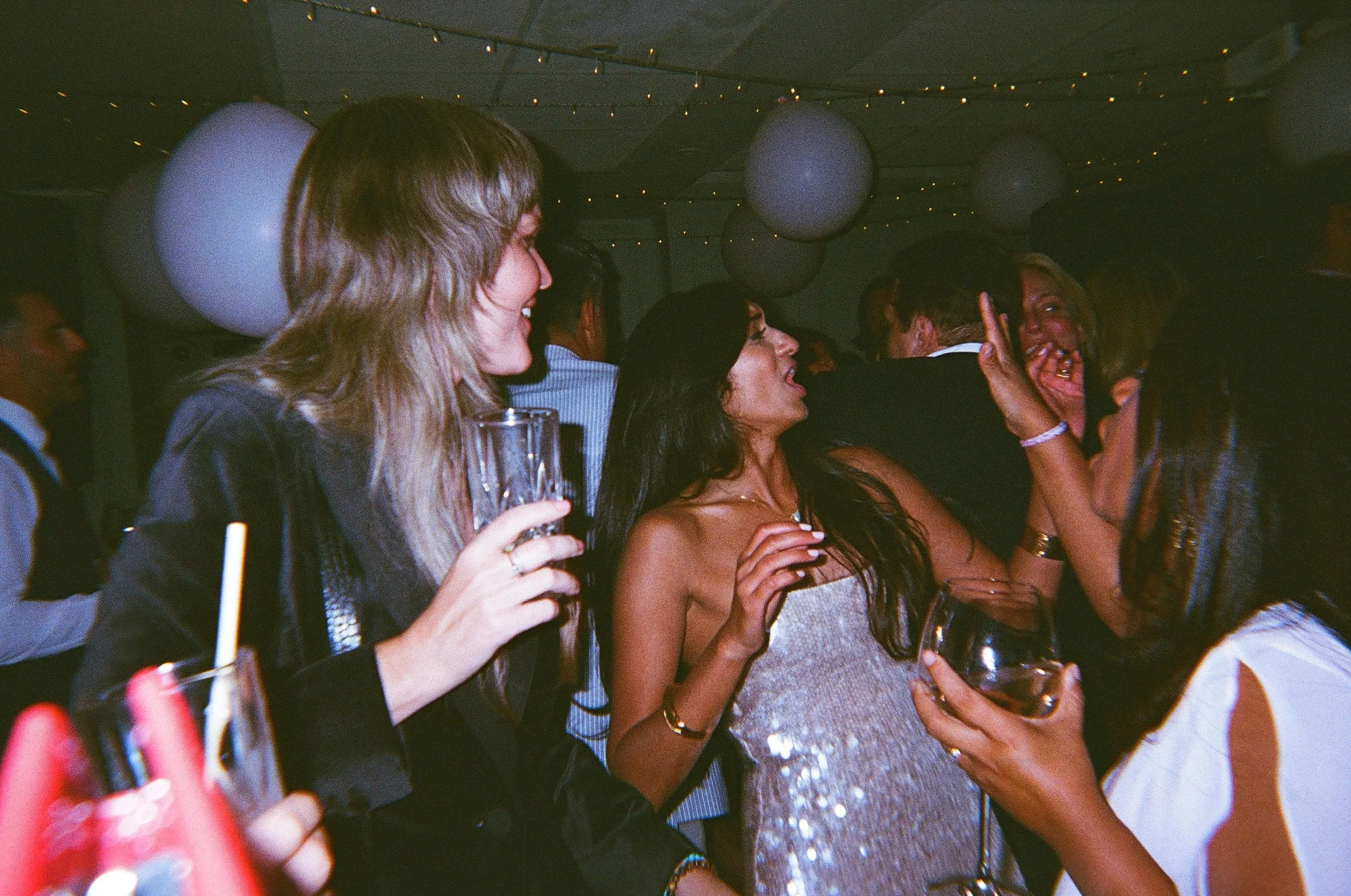 Group of friends enjoying a wedding party with drinks and dancing, party lights in the background.