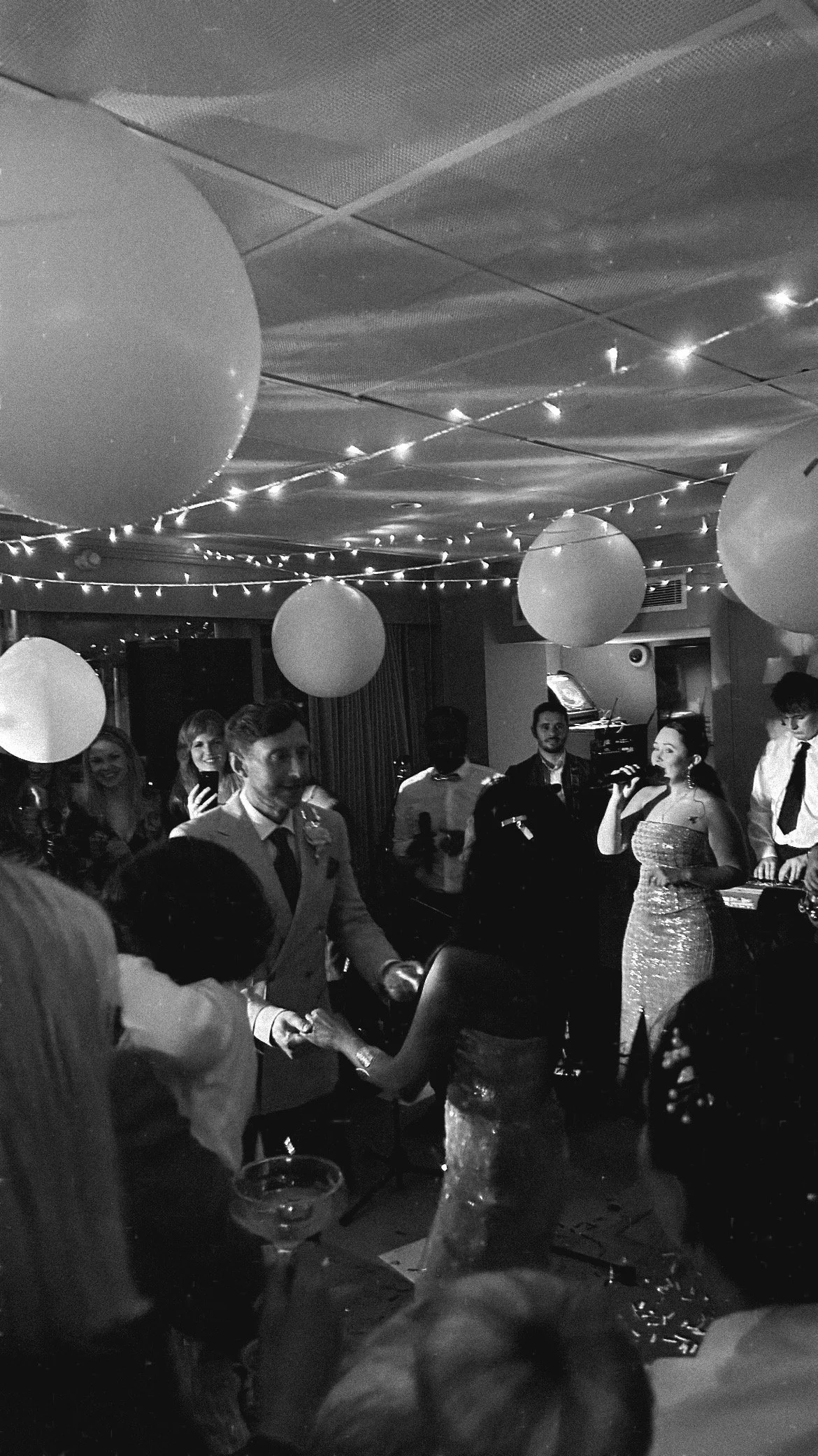 A black and white photo of a wedding reception with the newlyweds dancing, surrounded by friends and family, decorated with large balloons and string lights on the ceiling.