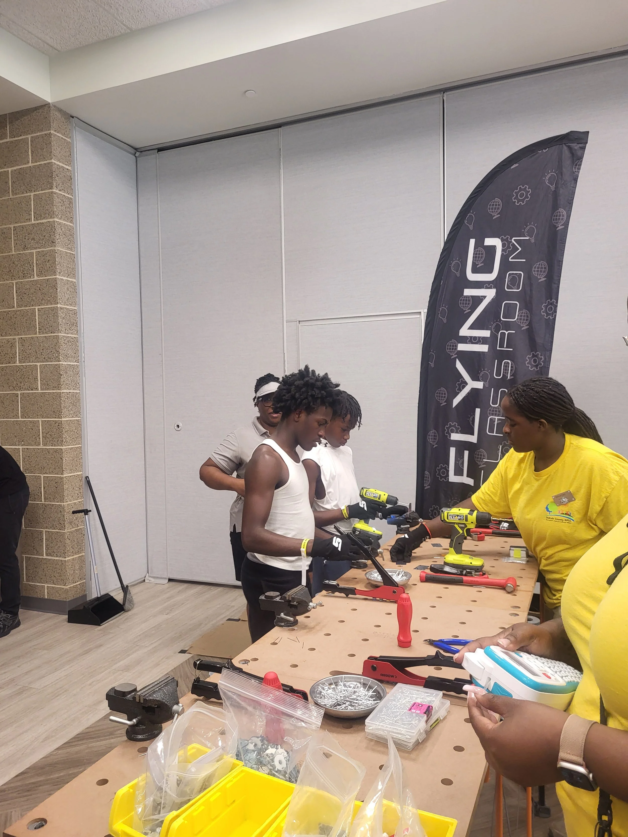Two young participants wearing work gloves use drills and hand tools at a Flying Classroom workbench while Commissioner Chakira Johnson and volunteers look on.