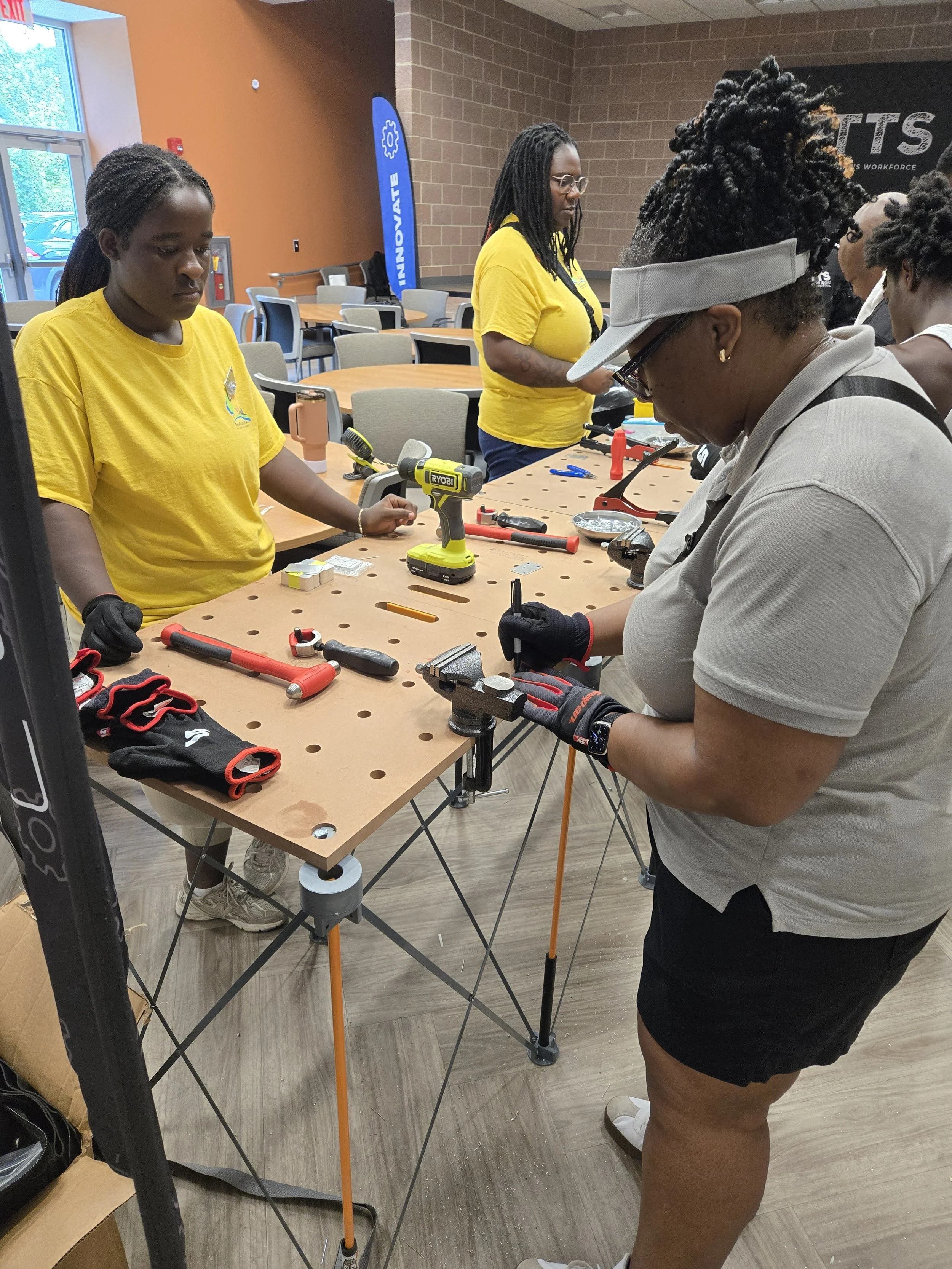 Commissioner Chakira Johnson, wearing work gloves, uses a hand tool on a metal piece secured in a table vise while a volunteer watches.