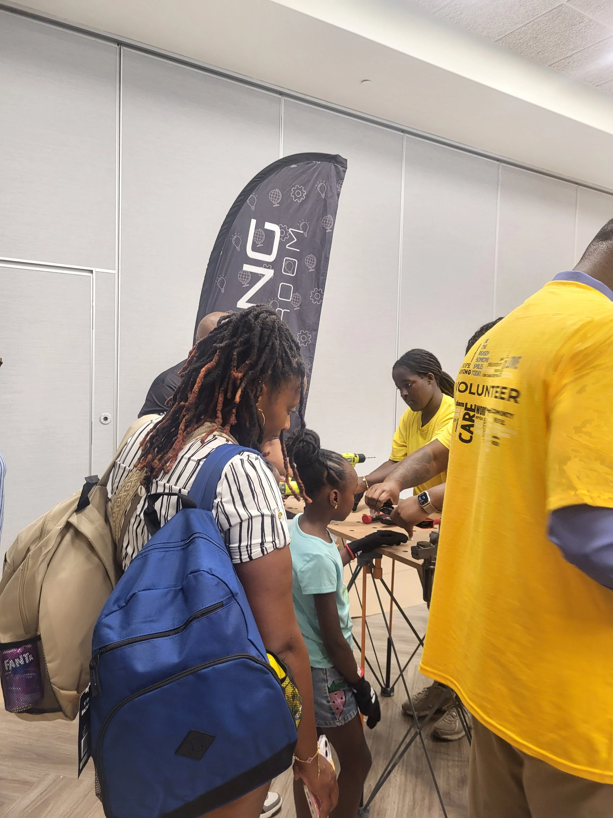 A child wearing safety gloves watches an instructor demonstrate a tool at a Flying Classroom activity station as family members and volunteers gather around.